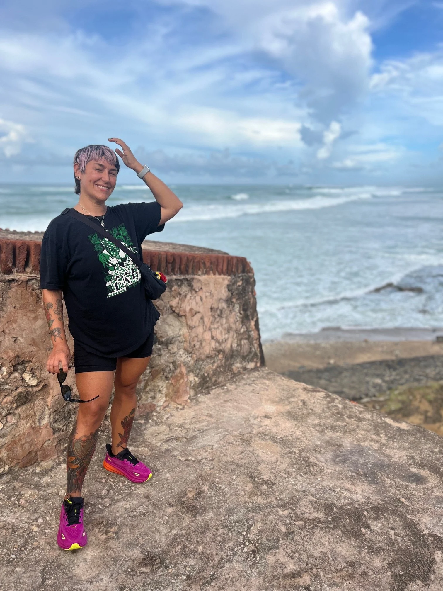 Woman with tattoos on legs and arm, wearing black shirt and shorts, smiling with eyes closed, standing on a rocky surface by the ocean with waves and partly cloudy sky in the background.