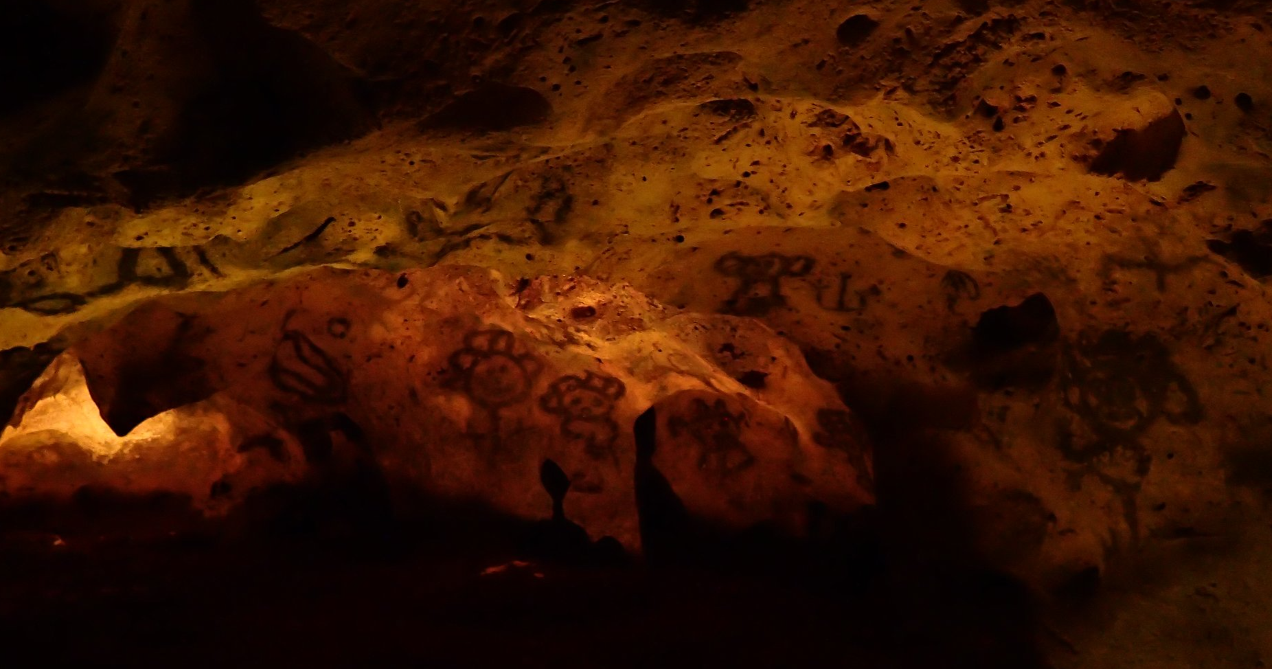 Dark cave wall with various graffiti, including drawings of owls, a robot, and the word 'PEACE' written with dark markings.