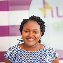 Smiling woman with short curly hair wearing a blue geometric patterned top, standing in front of a purple and white background with colorful signage.