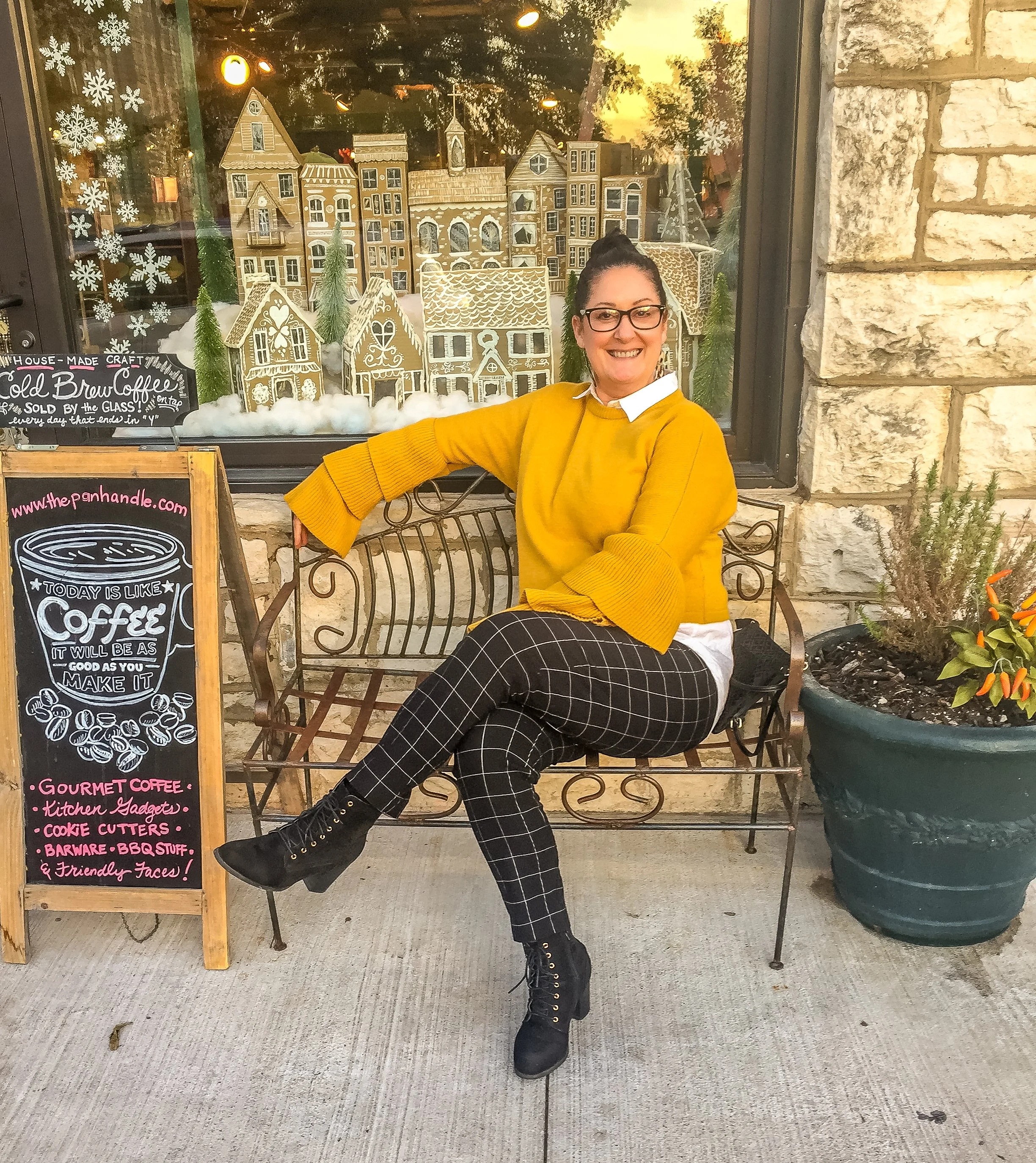 Woman with dark hair in a bun, wearing glasses, a yellow sweater, and black and white checkered pants, sitting on a metal bench outside a coffee shop, smiling at the camera. Behind her is a window decorated with gingerbread houses, snowflakes, green pine branches, and a sunset sky. To her left, there is a chalkboard sign advertising specialty coffee and baked goods.