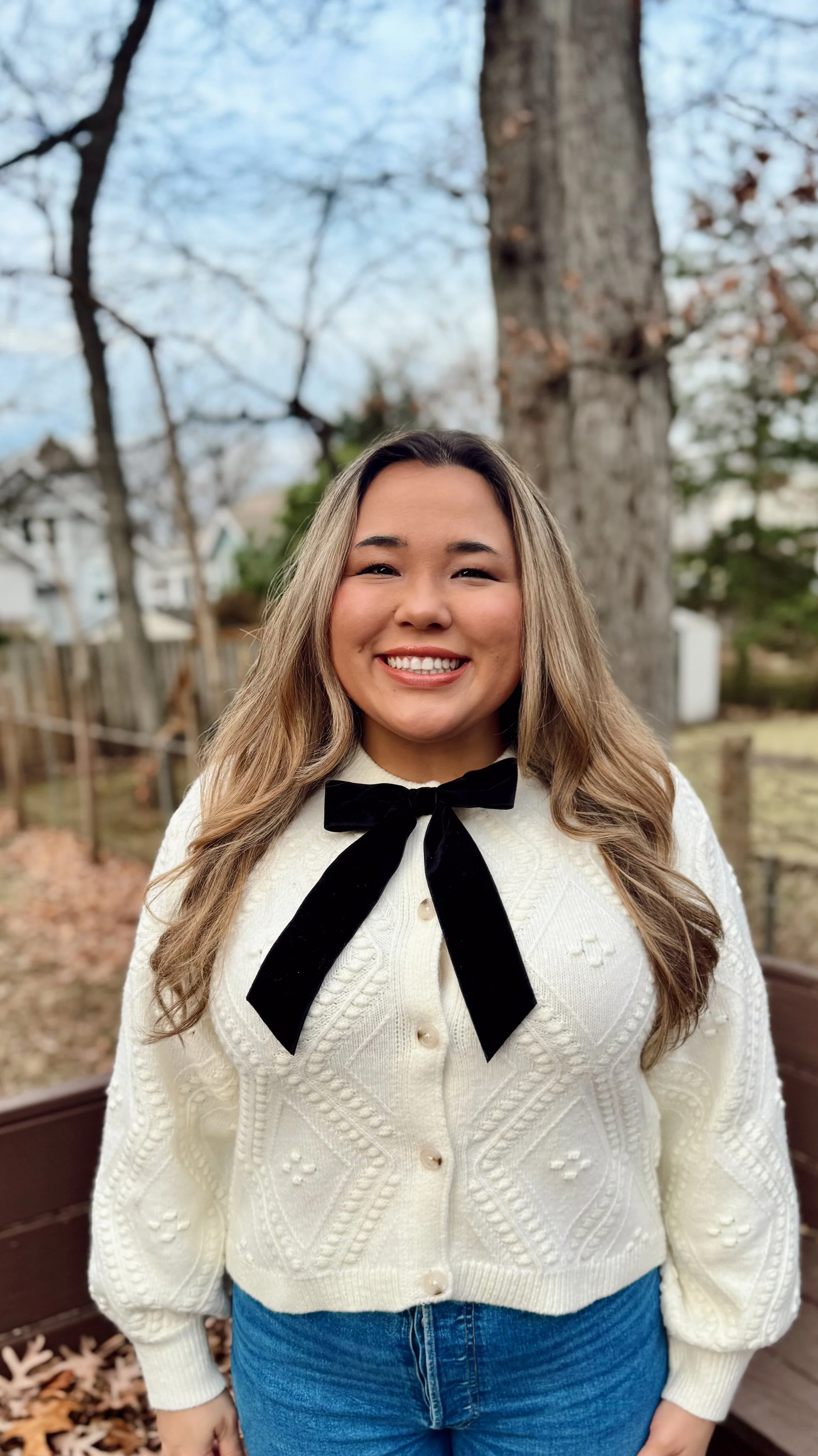 A smiling woman standing outdoors in front of a large tree with bare branches, wearing a cream cable-knit sweater with a black bow tie and blue jeans.