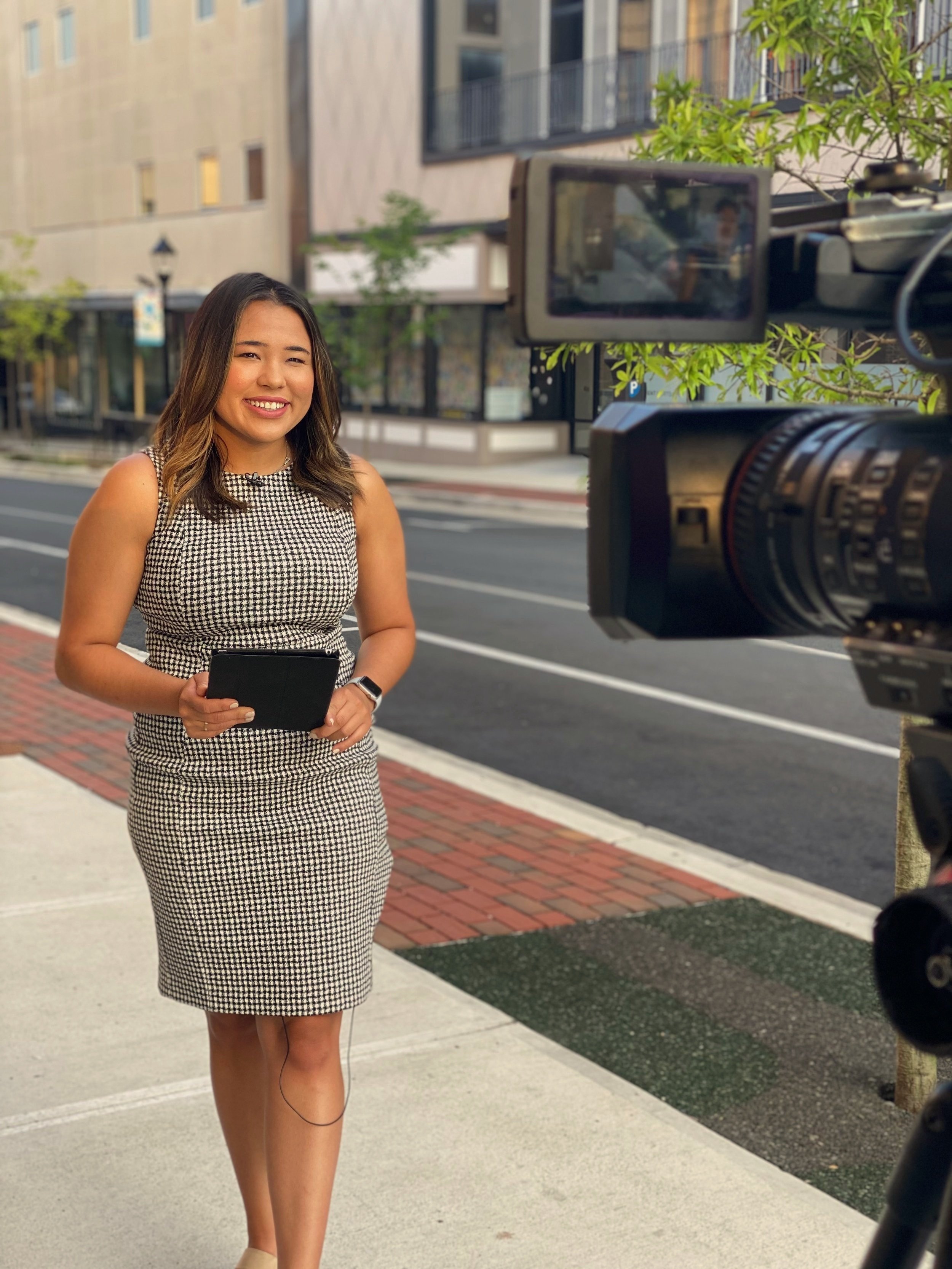 A woman being filmed on a city sidewalk with a professional video camera. She is smiling, wearing a black and white patterned dress, holding a tablet, and has a smartwatch on her wrist. There are buildings and trees in the background.