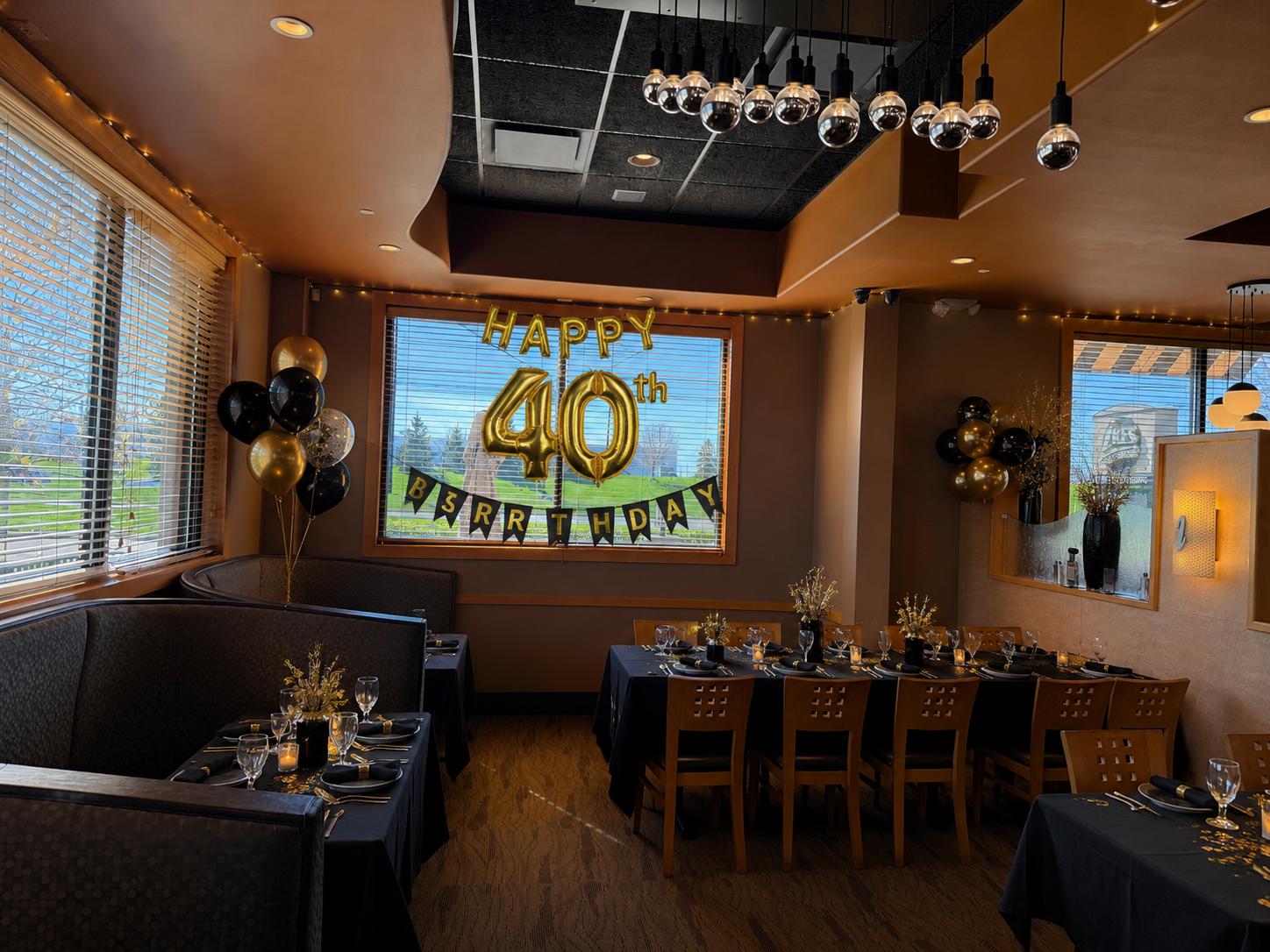 Decorated celebration room with black and gold balloons, a sign reading 'Happy 40th Birthday' in large gold balloons, a table set for a party with black tablecloths, wine glasses, candles, and floral centerpieces, and windows showing a sunny outdoor 