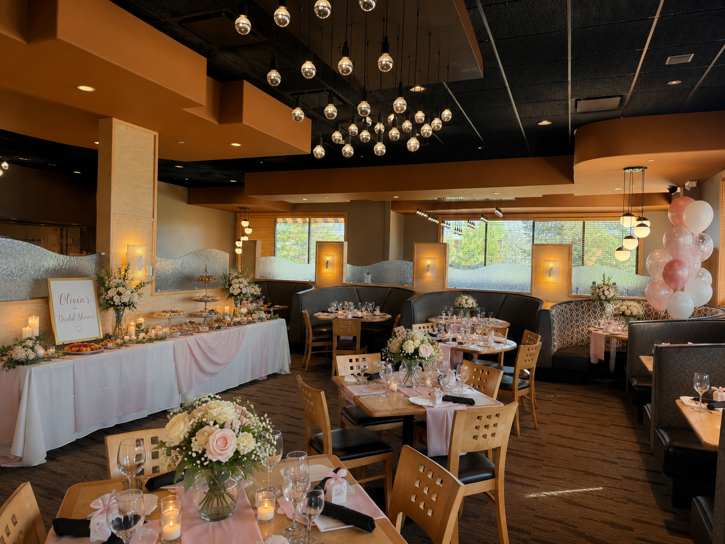 Elegant restaurant decorated for a bridal shower with pastel pink and white floral arrangements, balloons, and a dessert table, featuring round tables set with wine glasses, candles, and napkins.