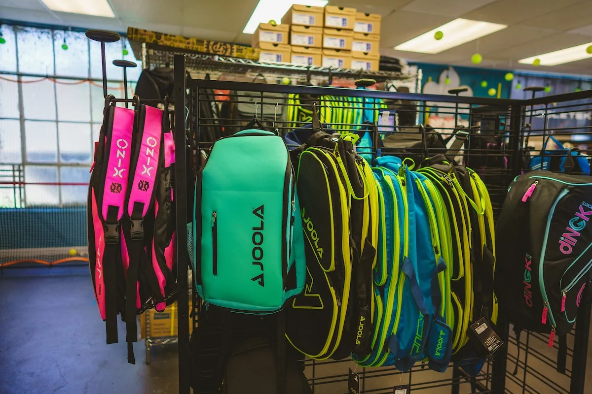 Colorful tennis bags hanging on a rack in a sporting goods store with a tennis court visible in the background.