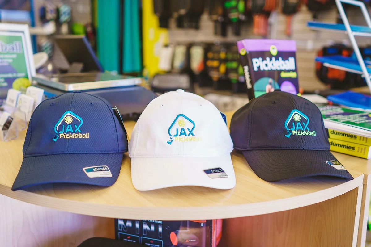 Three JAX Pickleball hats in navy blue, white, and black displayed on a store counter.