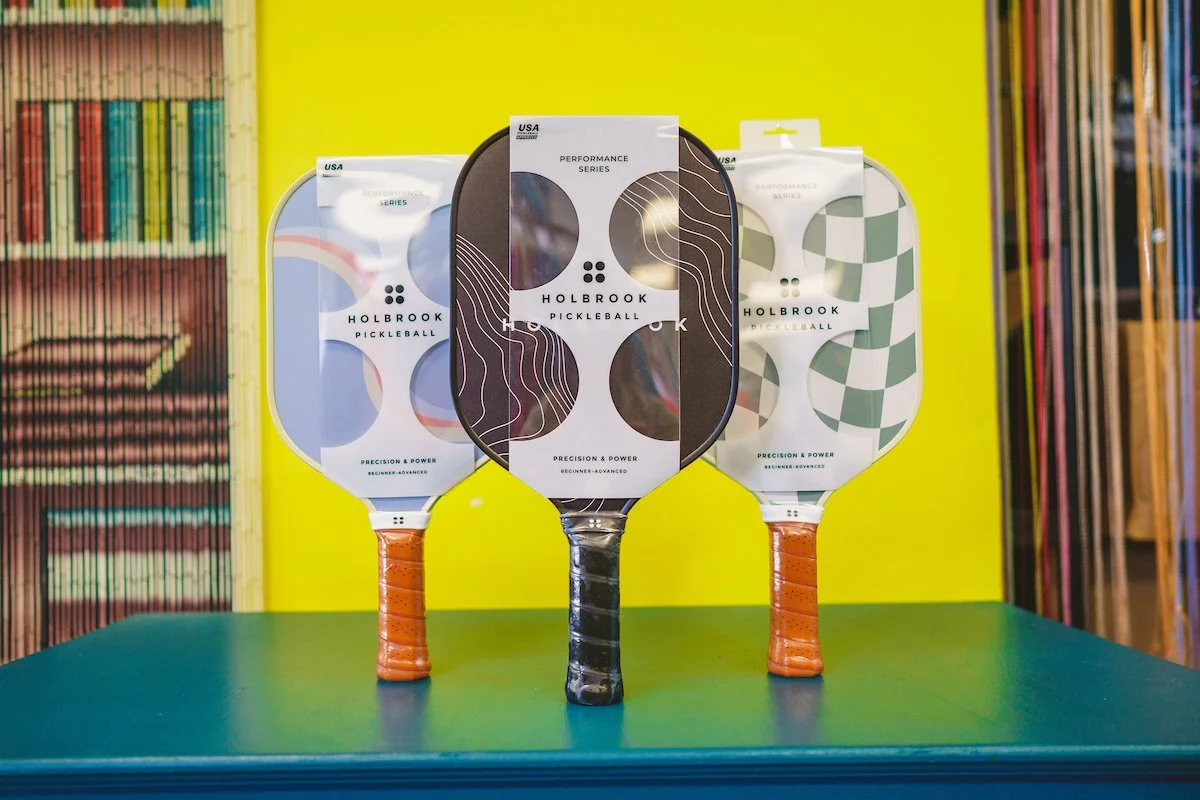 Three pickleball paddles standing upright on a table, with a yellow wall and various shelves in the background. The paddles have different designs on their faces.