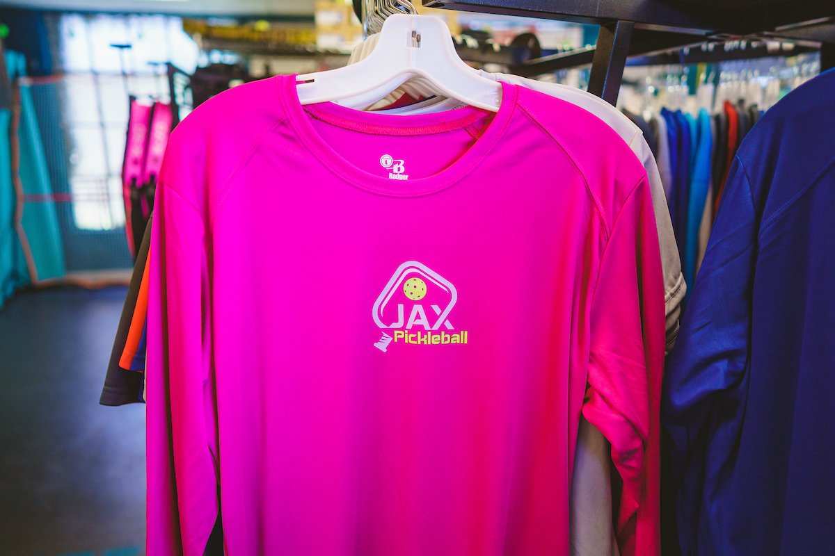 Pink long-sleeve athletic shirt with a JAX Pickleball logo on the front, hanging on a white hanger in a sports store.