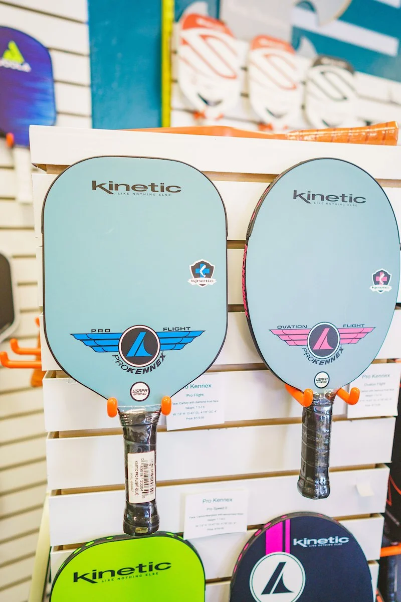 Four Kinetic brand pickleball paddles displayed on a store rack in a sporting goods store, with colorful paddles visible in the background.