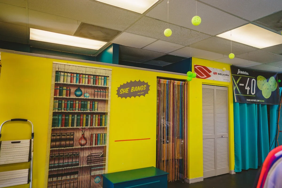 Colorful indoor space with yellow walls, a blue door with a multicolored beaded curtain, and hanging green balls from the ceiling. A bookshelf mural and a sign that says 'She Bangs' are visible. There are also banners and a blue curtain on the right.
