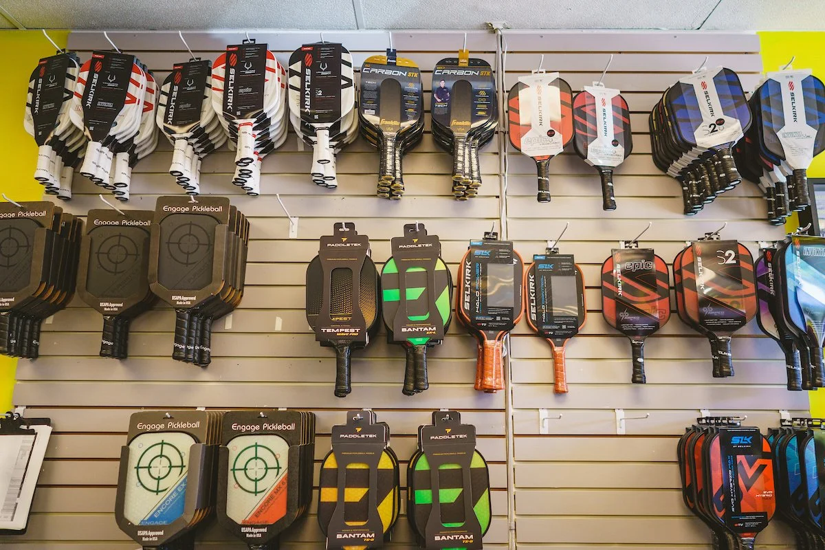 A display of pickleball paddles hanging on a slatwall, arranged in rows by style and brand.