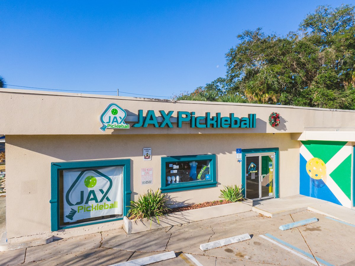 Exterior of JAX Pickleball sports store with signage, window display, parking spaces, and a mural, surrounded by trees and under a clear blue sky.