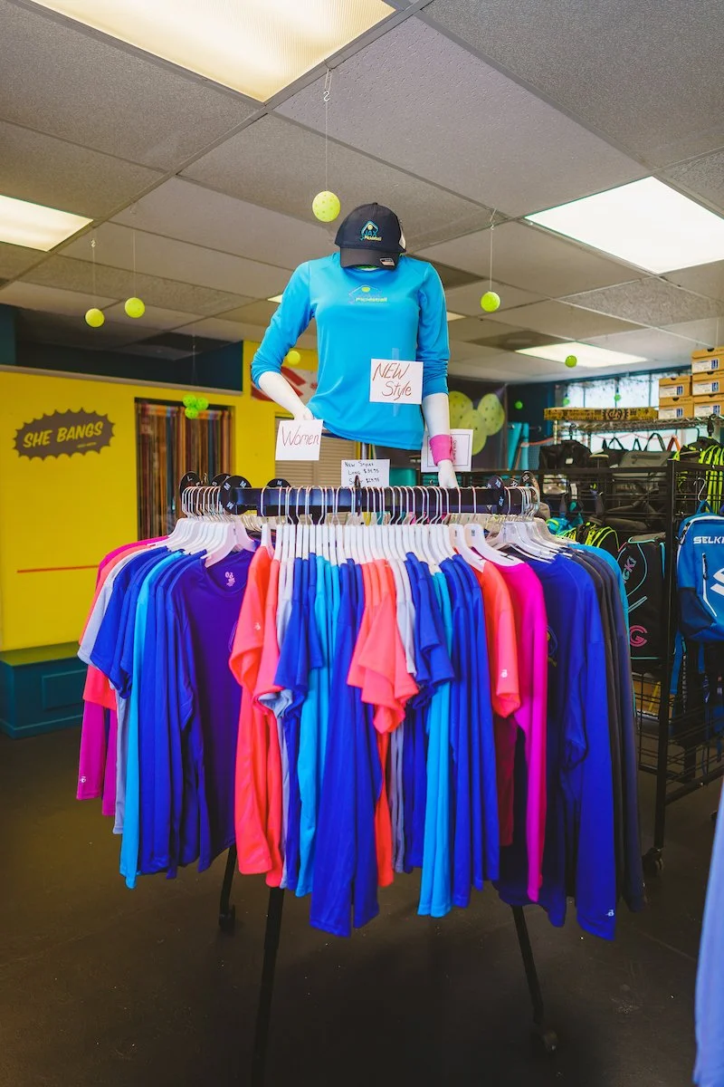 A clothing display featuring colorful athletic shirts hung on a rack in a retail store, with a mannequin wearing a blue shirt and a cap on top. Signs indicate new styles and women’s apparel, and tennis balls are hanging from the ceiling as decor.