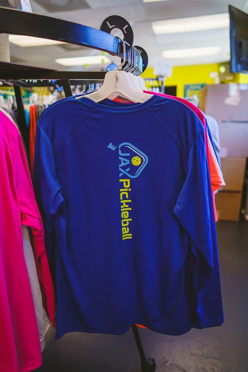 Blue athletic shirt with the text 'JAX Pickleball' and a pickleball paddle logo hanging on a rack in a store.