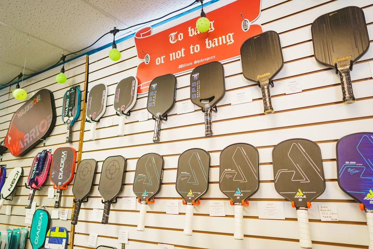 A display of pickleball paddles hanging on a slatwall, with a red banner that has a quote by William Shakespeare, "To be or not to be."