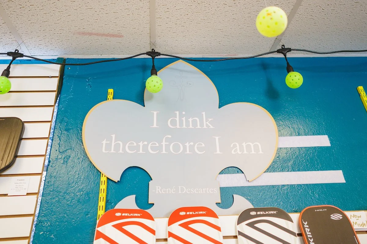 A wall display with a quote from René Descartes saying, "I think therefore I am," with puzzle piece shapes and skateboards below, in a retail or skate shop setting. Green string lights are hanging above.