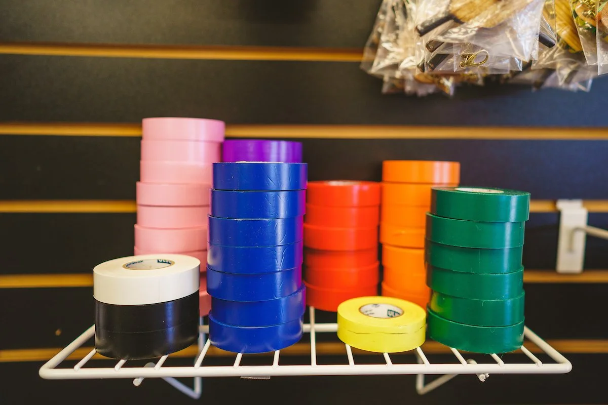 Stacks of colorful adhesive tape rolls in pink, purple, blue, red, orange, green, yellow, and black on a white wire rack.