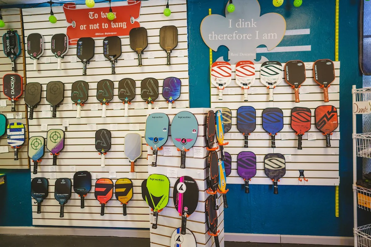 Display of pickleball paddles arranged on a wall at a sports store, with a sign that says "To bat or not to bat" and another sign shaped like a cloud reading "I dink therefore I am."
