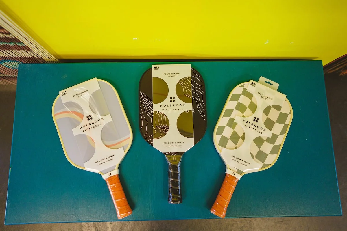 Three pickleball paddles on a teal surface, each with different designs and orange handles, branded with Holbrook Pickleball.