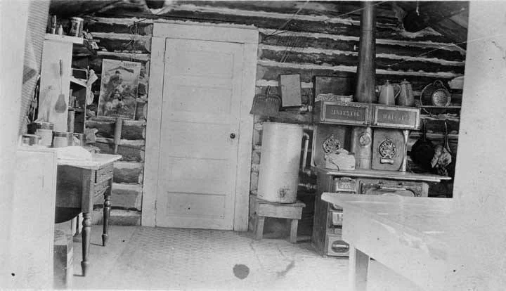 an old-time kitchen with a wood-fired cook stove, water can, and tables around a door in a chinked-log building