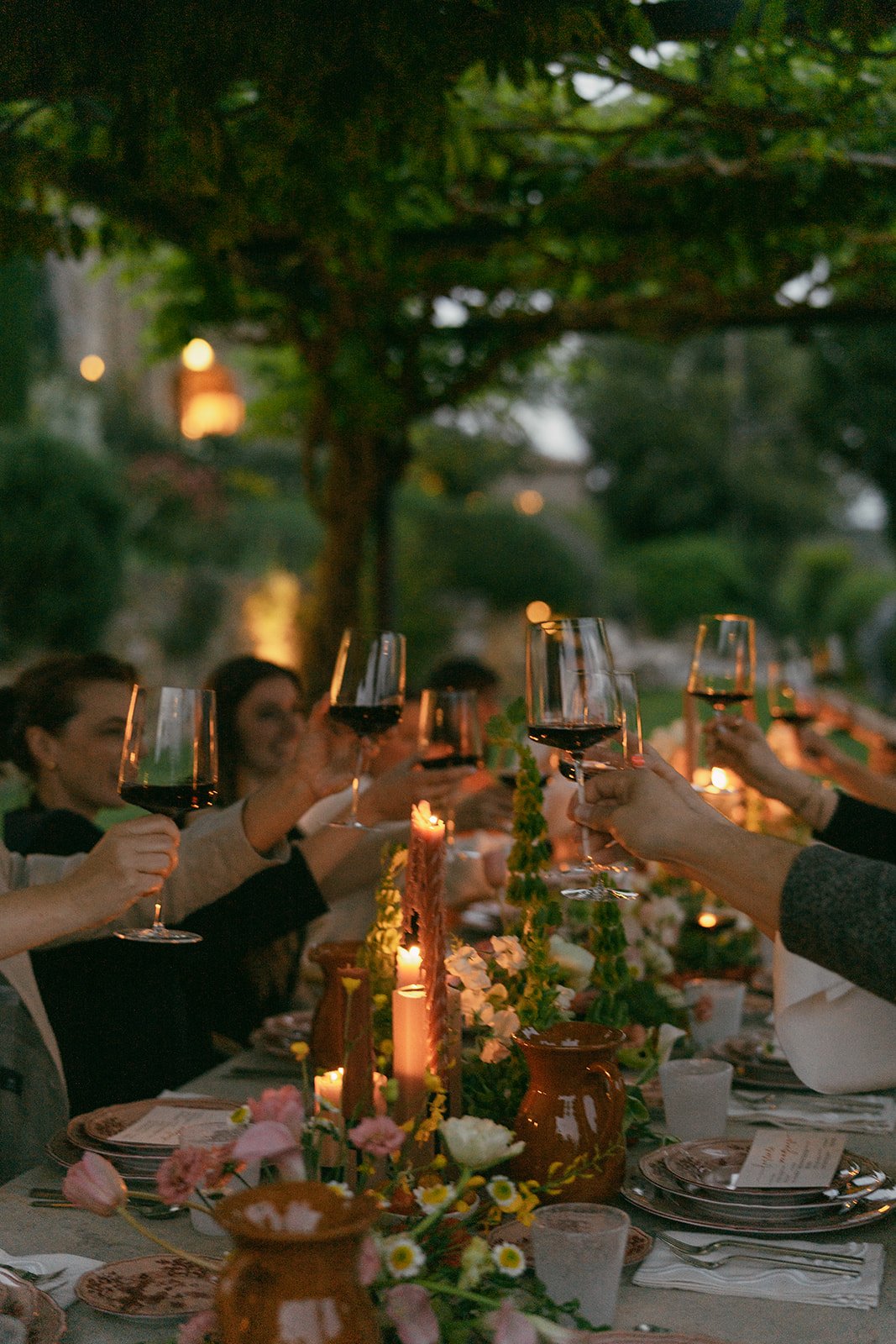Outdoor dinner party with people toasting with wine glasses, table decorated with candles and flowers.