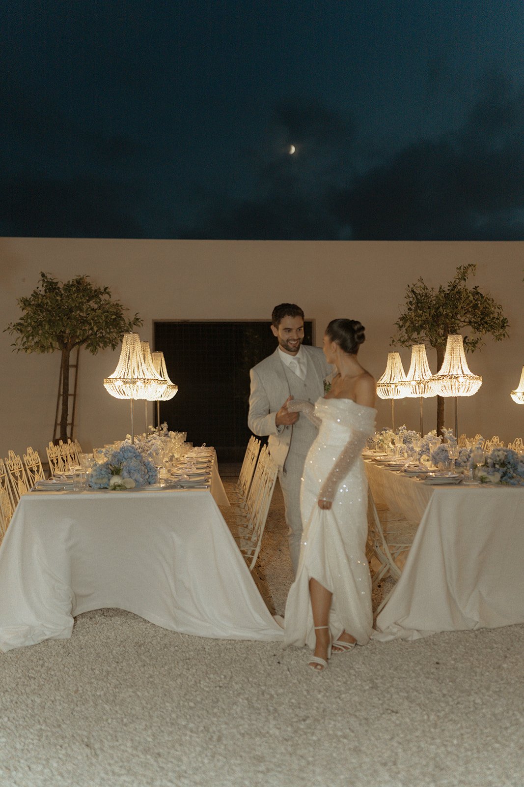 A couple dressed in formal attire in an outdoor evening wedding setting with chandelier lighting and elegantly set tables with white tablecloths and floral arrangements.