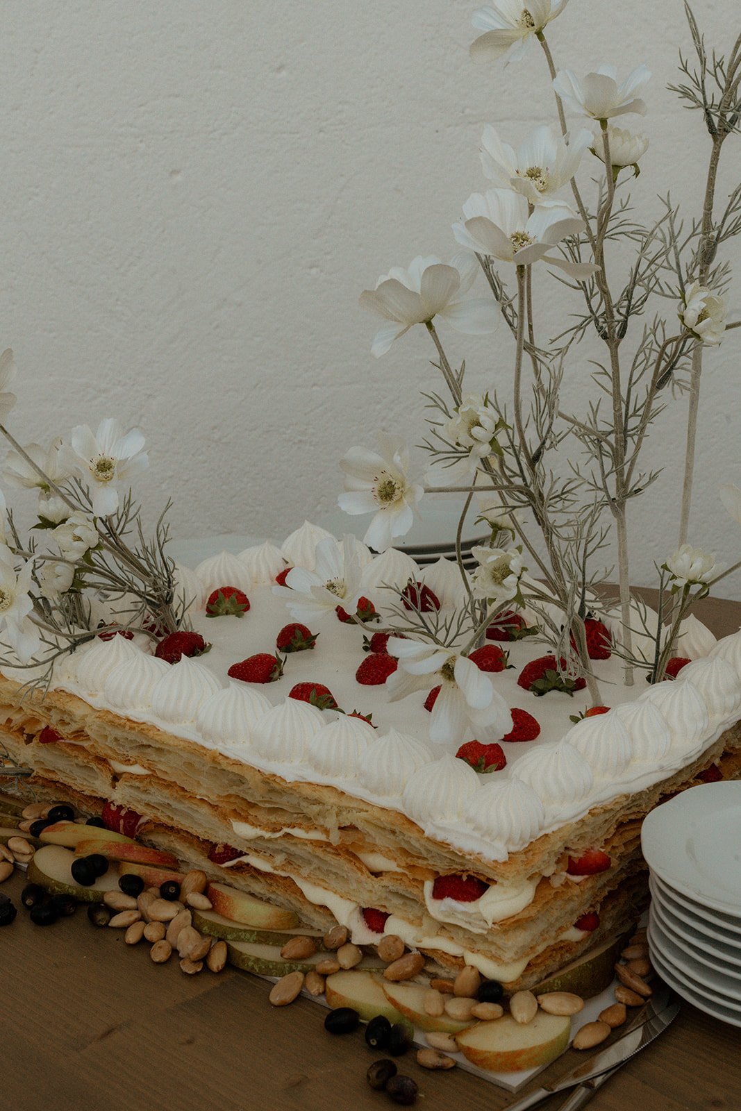 Layered cream cake with strawberries and decorative white flowers, surrounded by fruits, nuts, and empty plates.
