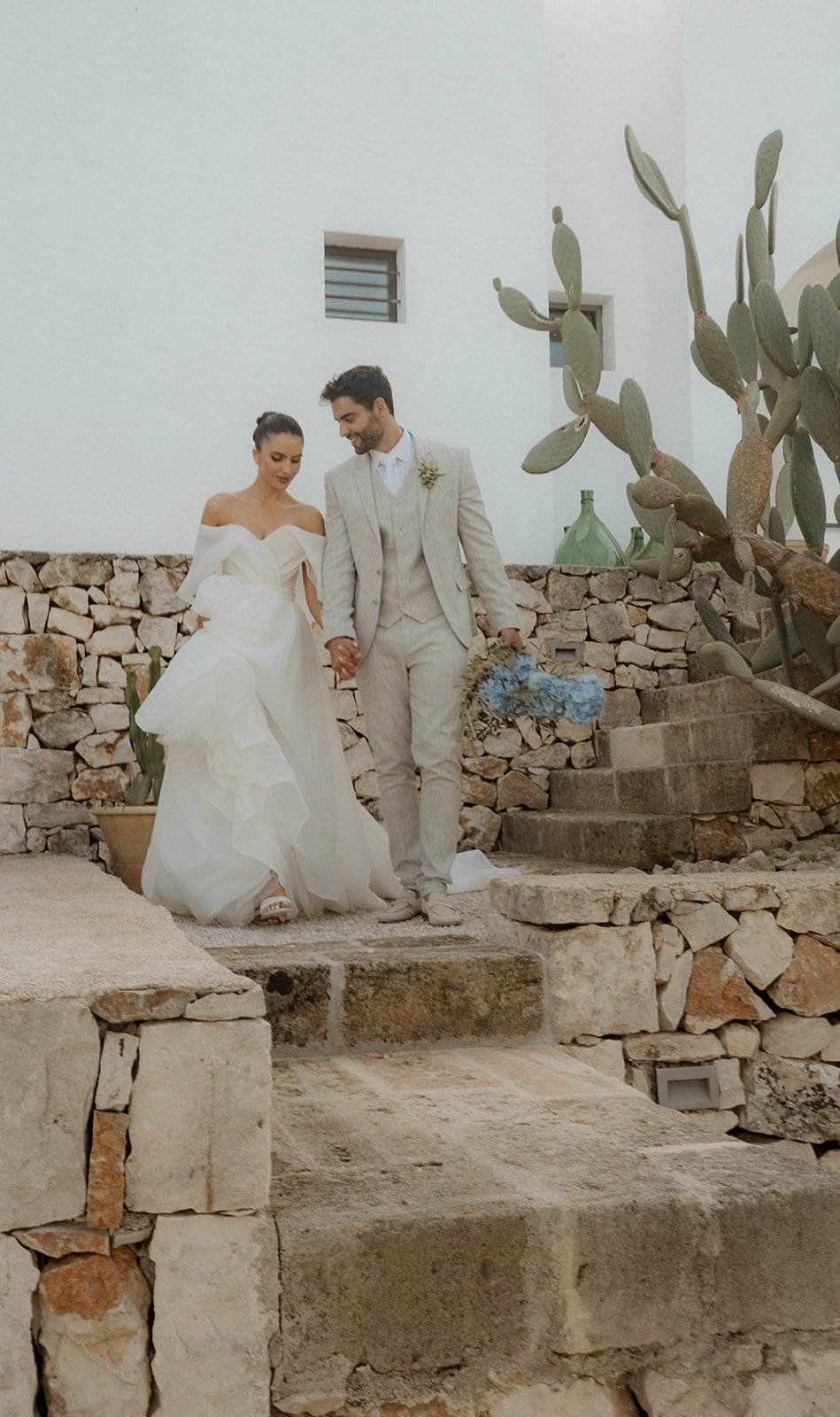 Bride and groom walking hand in hand on stone steps, with a cactus and white building in the background. The bride wears an off-shoulder wedding dress; the groom wears a light-colored suit.
