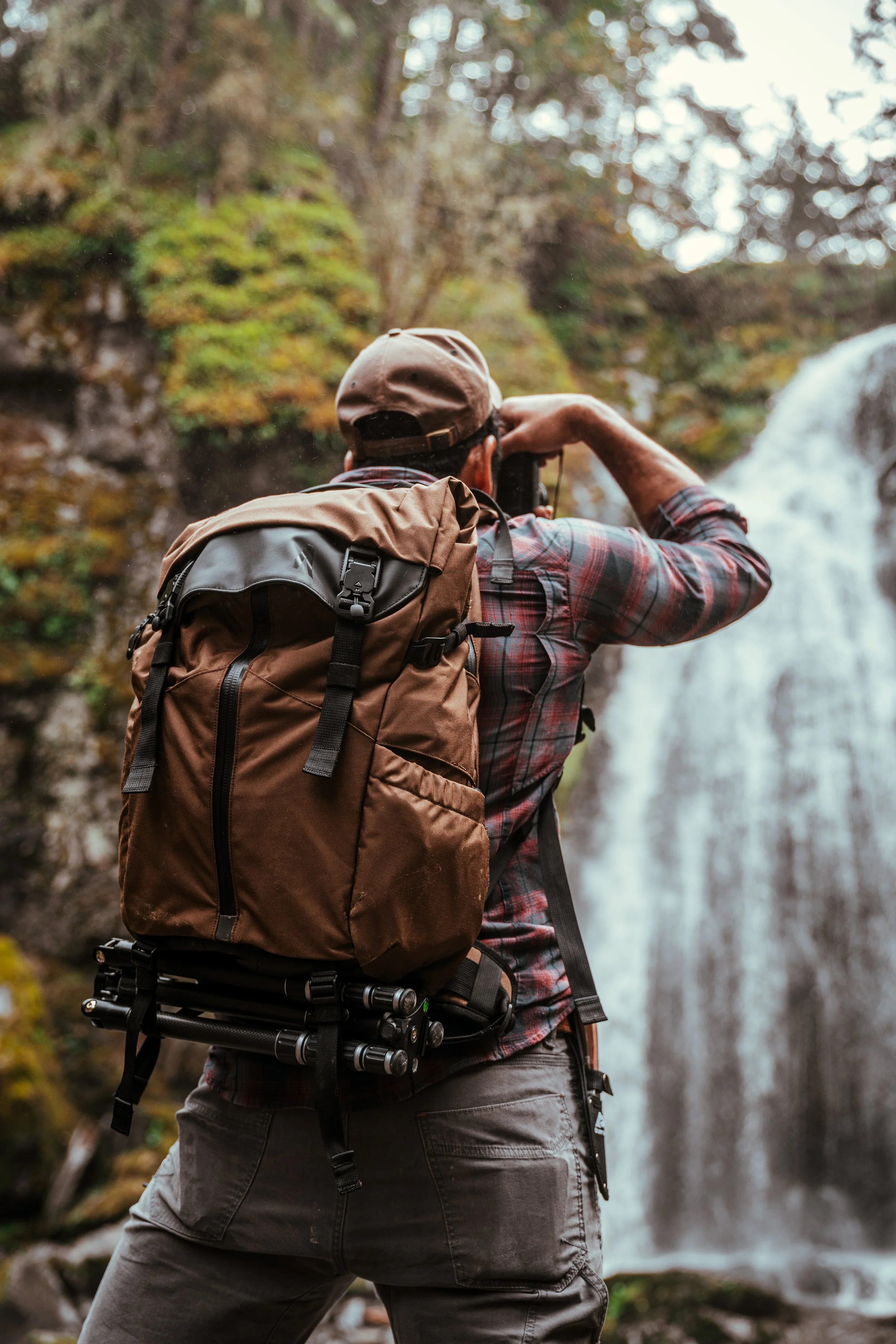 A man with a brown backpack and a cap taking photographs of a waterfall in a forested area.