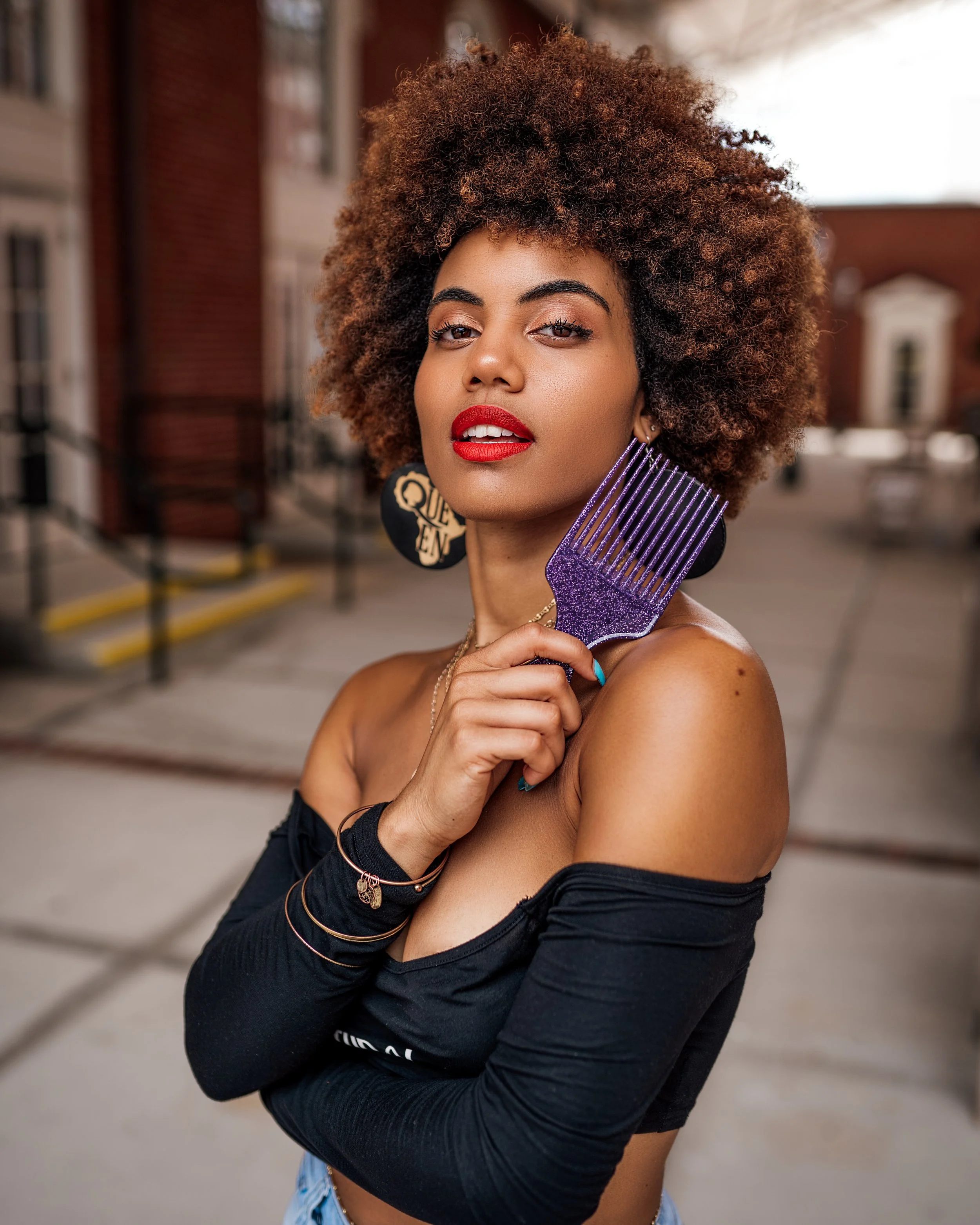 A woman with curly hair and red lipstick holding a purple comb at an outdoor urban location.