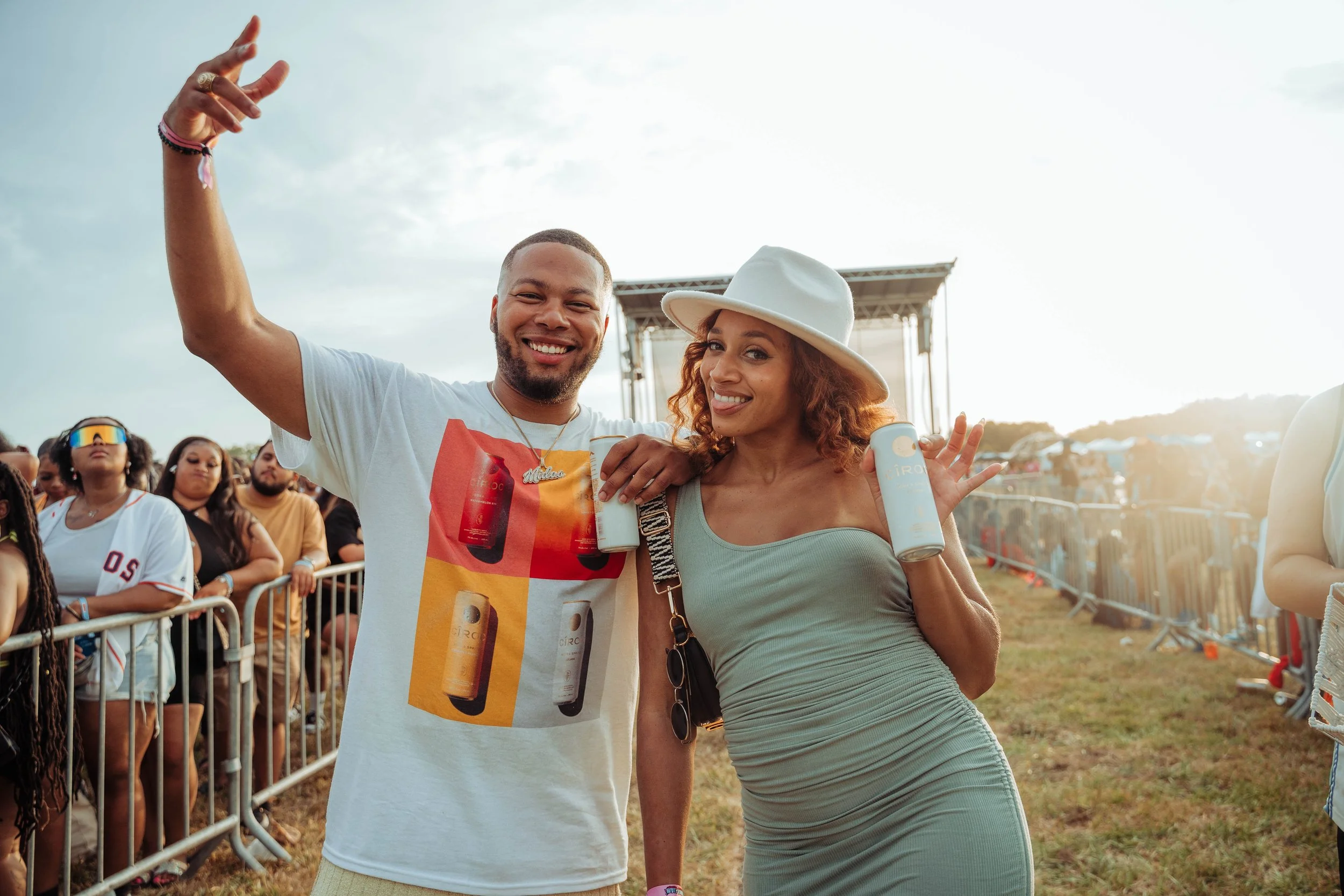 Two people smiling at an outdoor event, holding cans of drinks, with a crowd and stage in the background during sunset.