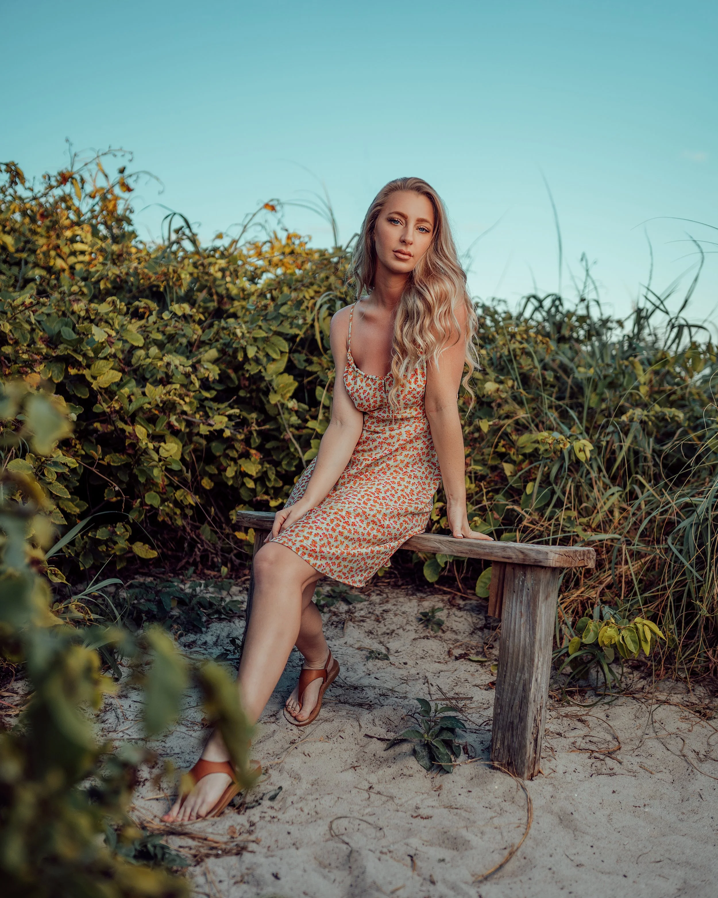 A woman with long blonde hair sitting on a wooden bench surrounded by green bushes and sand, wearing a floral dress and sandals on a beach or park setting.