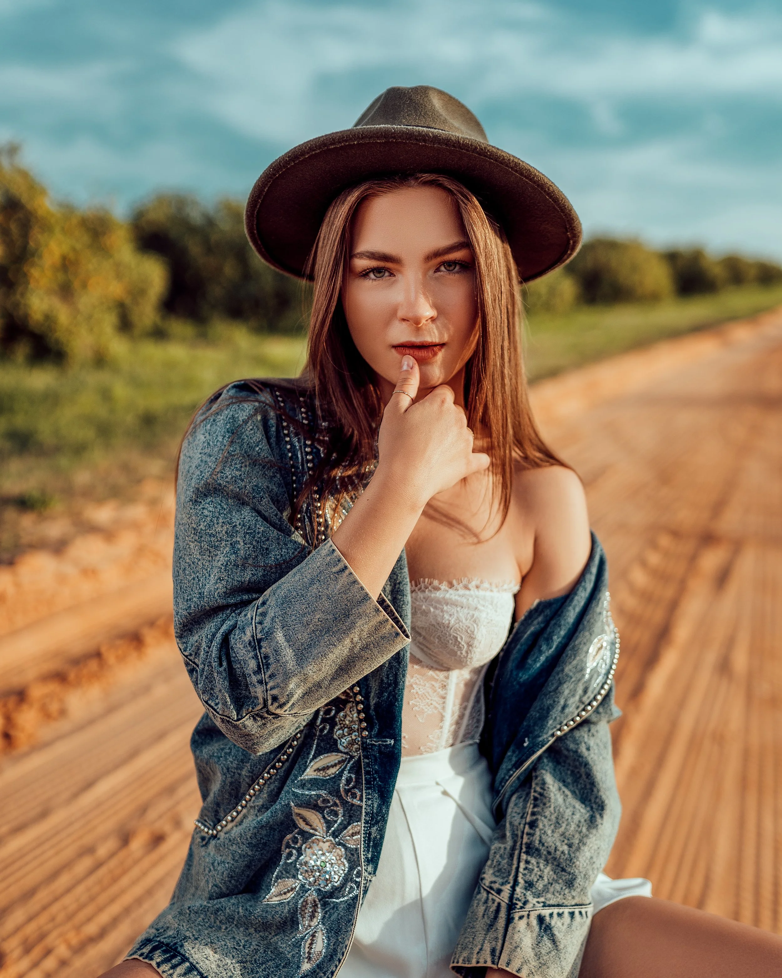 Young woman in a denim jacket, white lace top, and wide-brimmed hat sitting on a dirt path in a rural area with trees in the background.