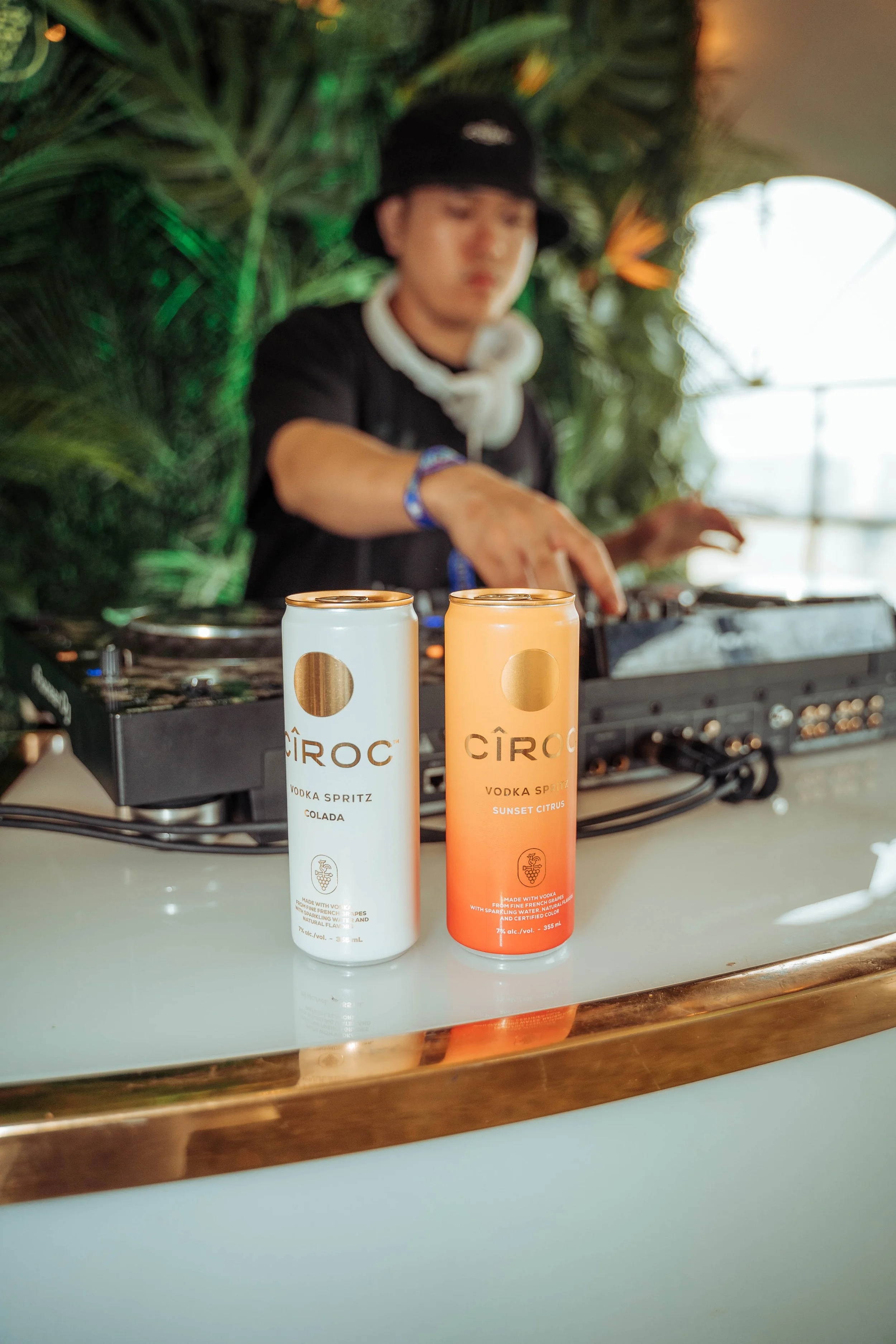Two cans of CÎROC vodka spritz drinks, one with white and purple packaging labeled 'Colada' and the other with orange and pink packaging labeled 'Sunset Citrus', placed on a bar counter with a DJ in the background and tropical plants.