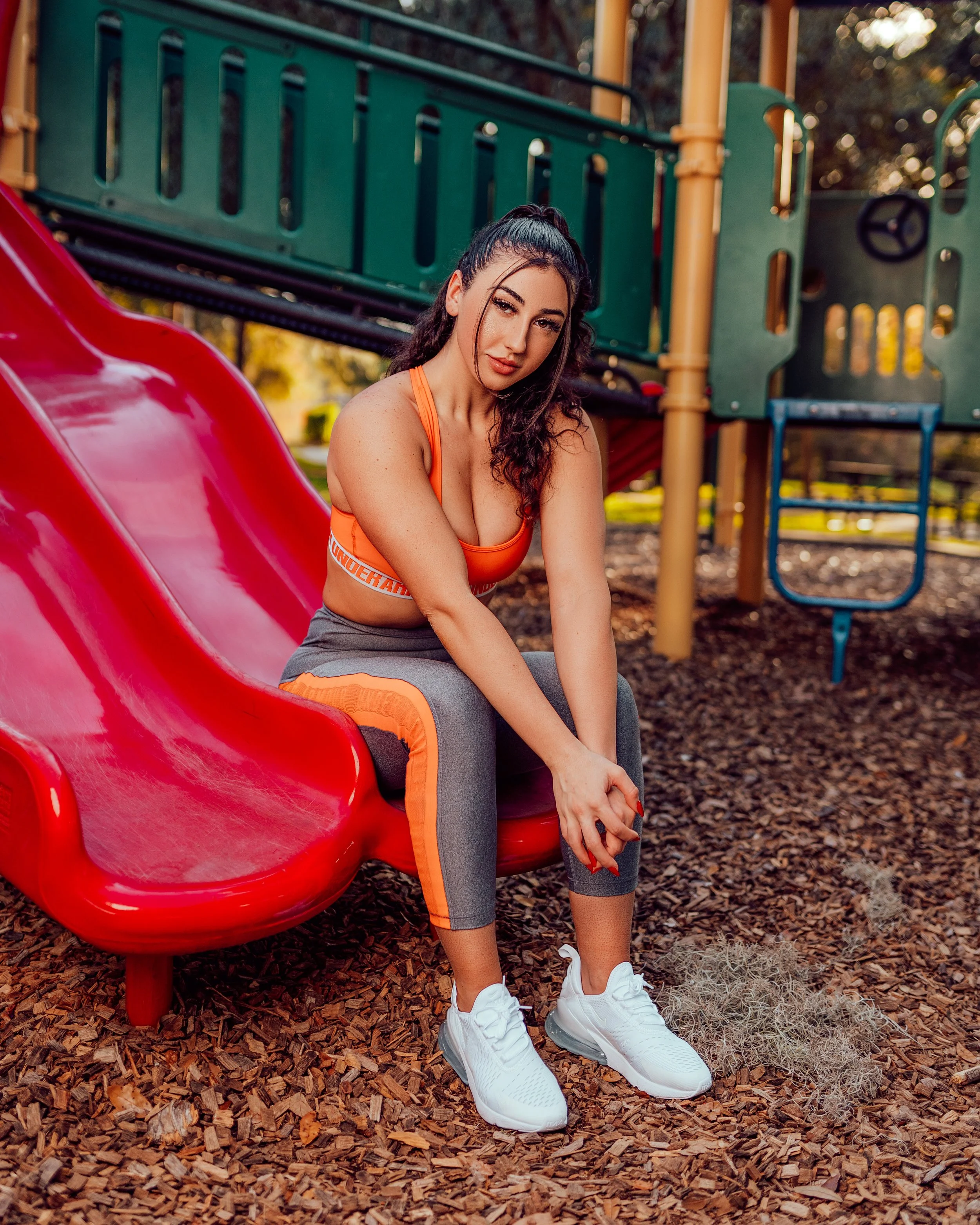 A woman wearing an orange sports bra and gray athletic pants with orange accents, sitting on a red slide at a playground, surrounded by wood chips, with outdoor play structures in the background.