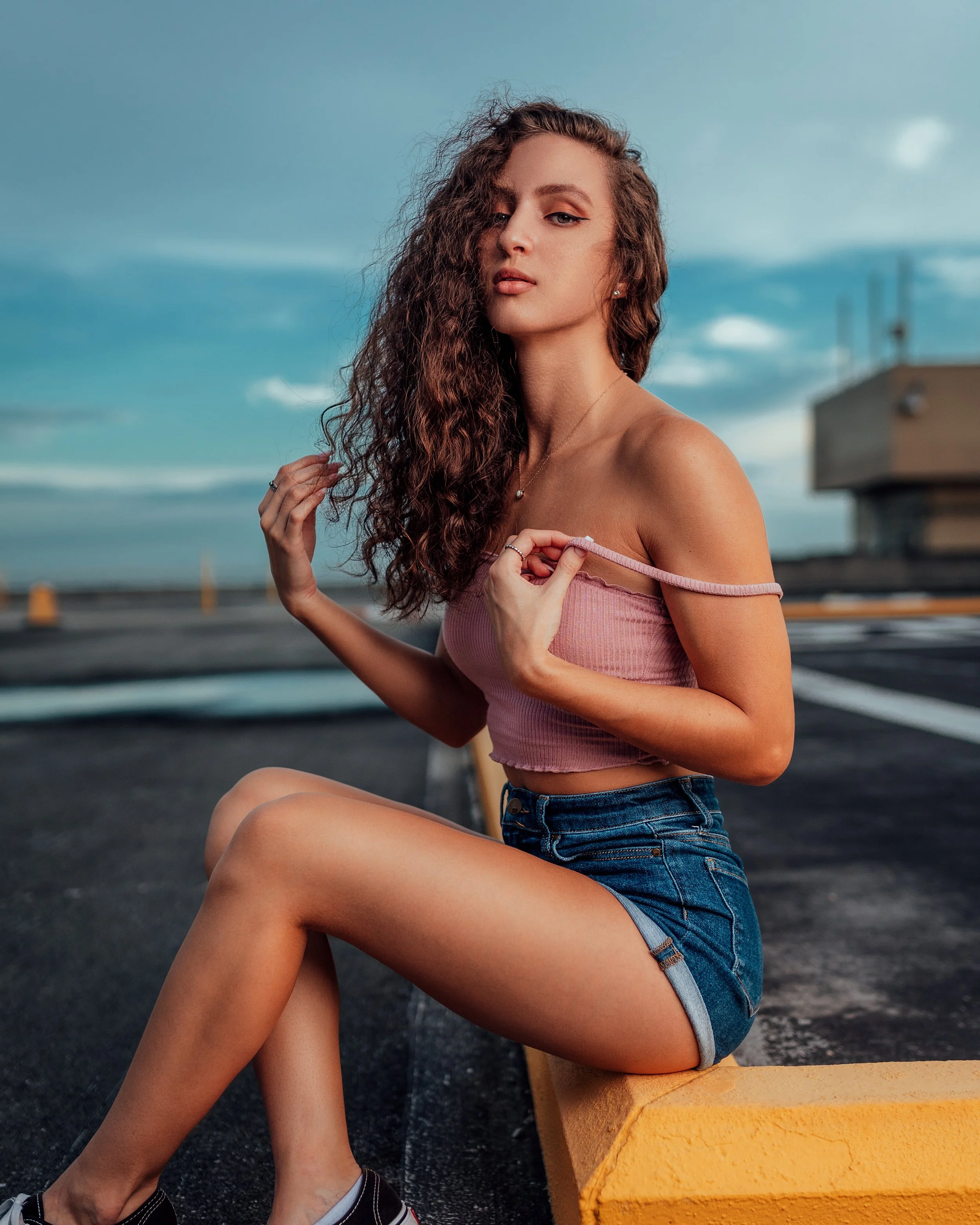 A young woman with long, curly brown hair sitting on a yellow parking block on the rooftop of a parking garage, wearing a pink strapless top and denim shorts, with an overcast sky in the background.