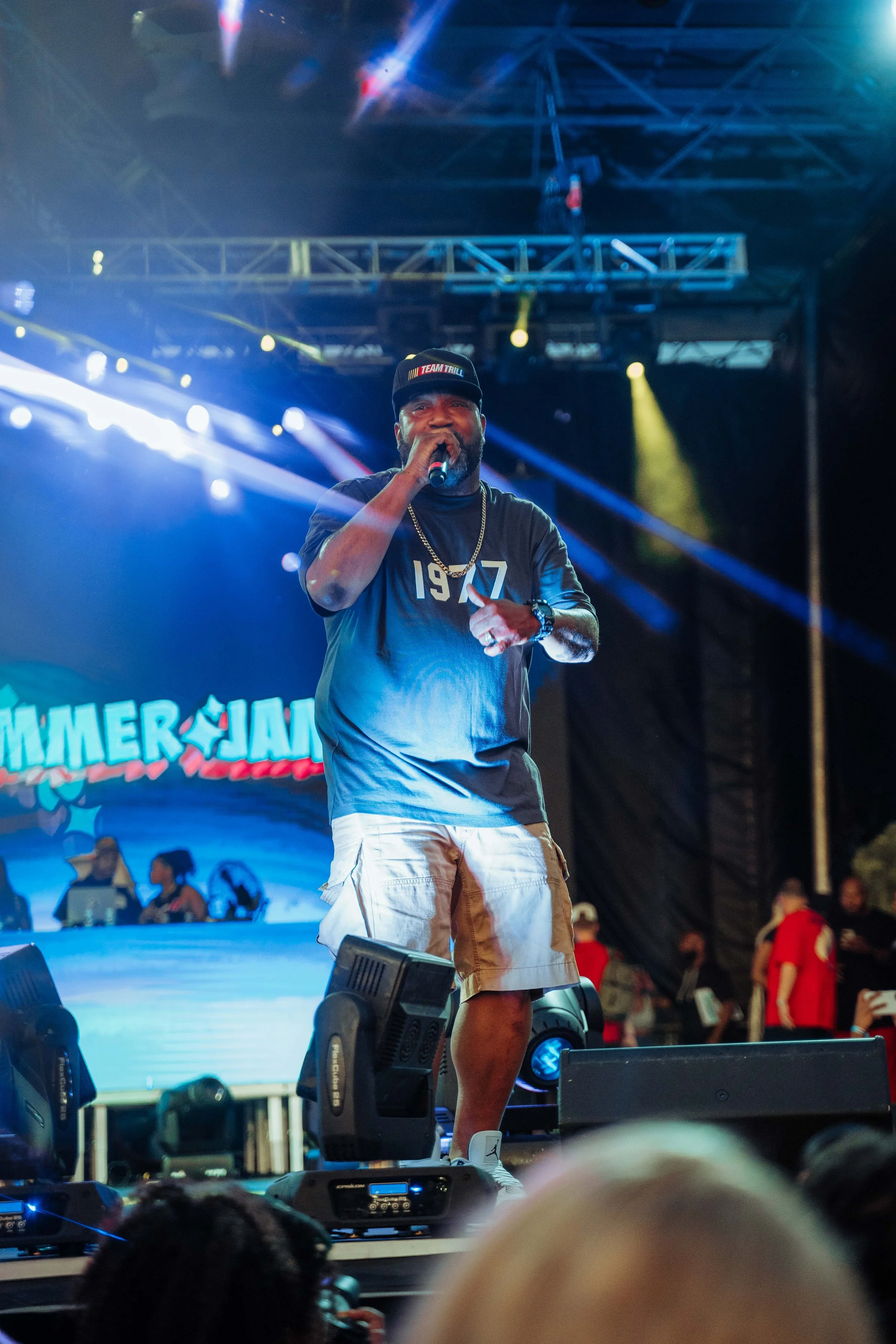 A man performs on stage at a concert, holding a microphone and wearing a black cap and t-shirt, with bright stage lights and a LED screen in the background.