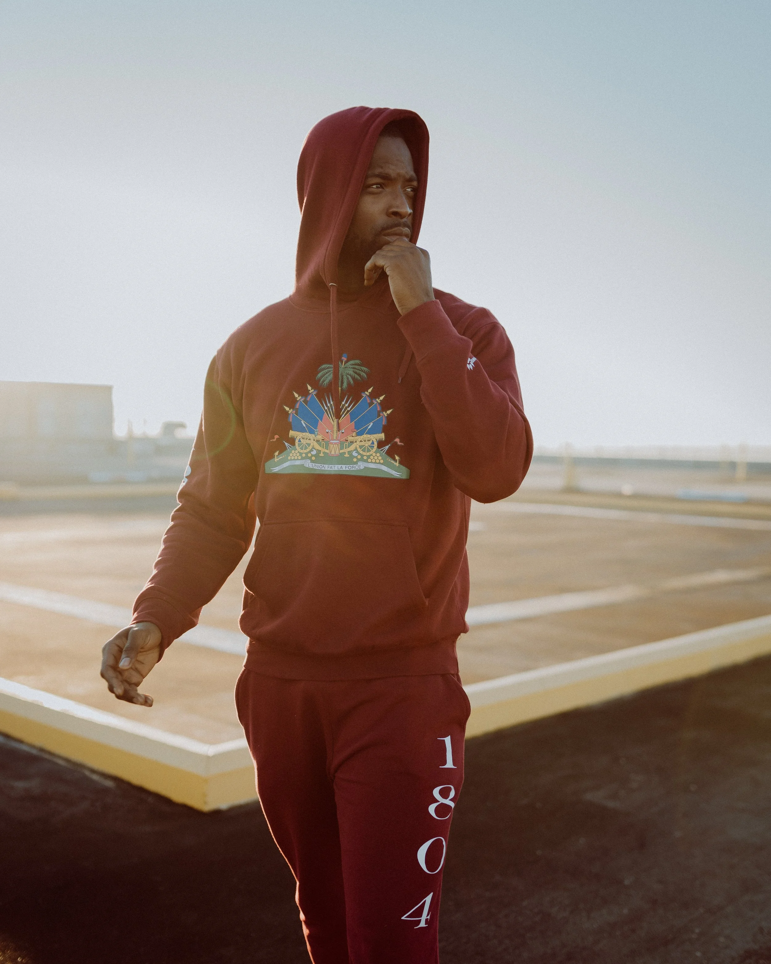 A man wearing a maroon hoodie with a colorful graphic on the front and maroon sweatpants with the year 1804 printed on the side, standing on a rooftop during daylight hours, with a thoughtful expression and hand near his chin.