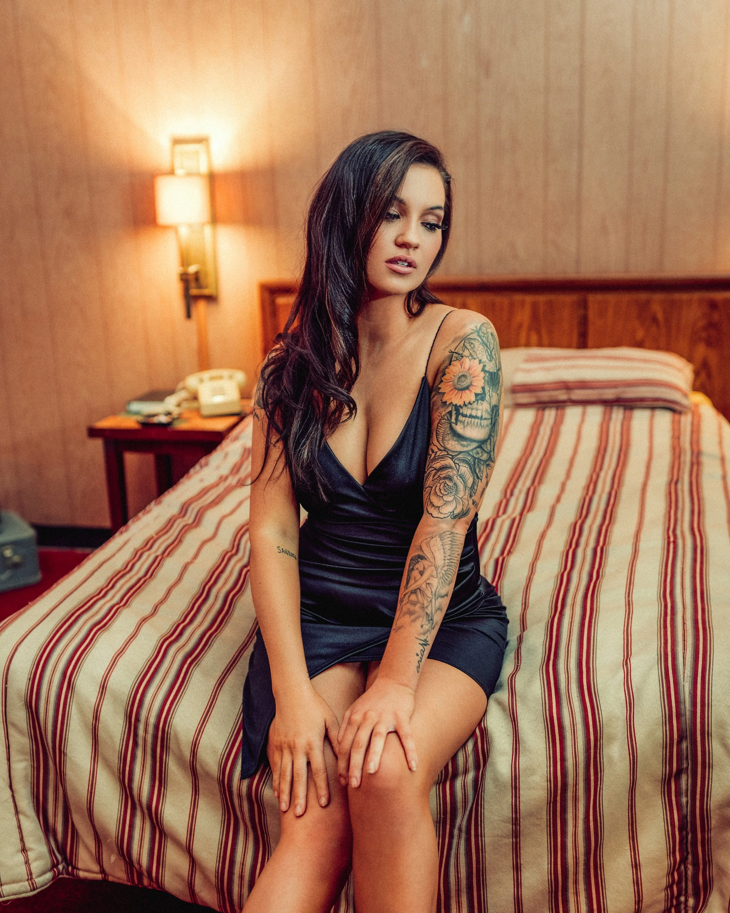A woman with dark hair and tattoos on her arms sitting on a bed in a hotel room, wearing a black silky dress.