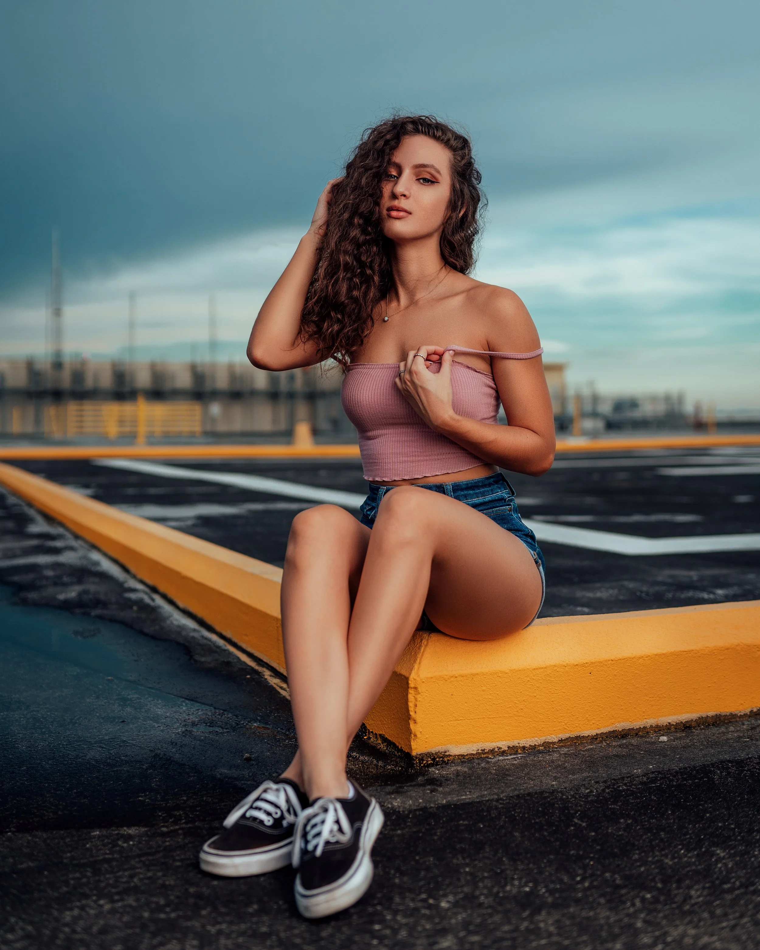Young woman with curly hair sitting on the edge of a parking lot curb, wearing a pink crop top, denim shorts, and black sneakers, with a cloudy sky in the background.