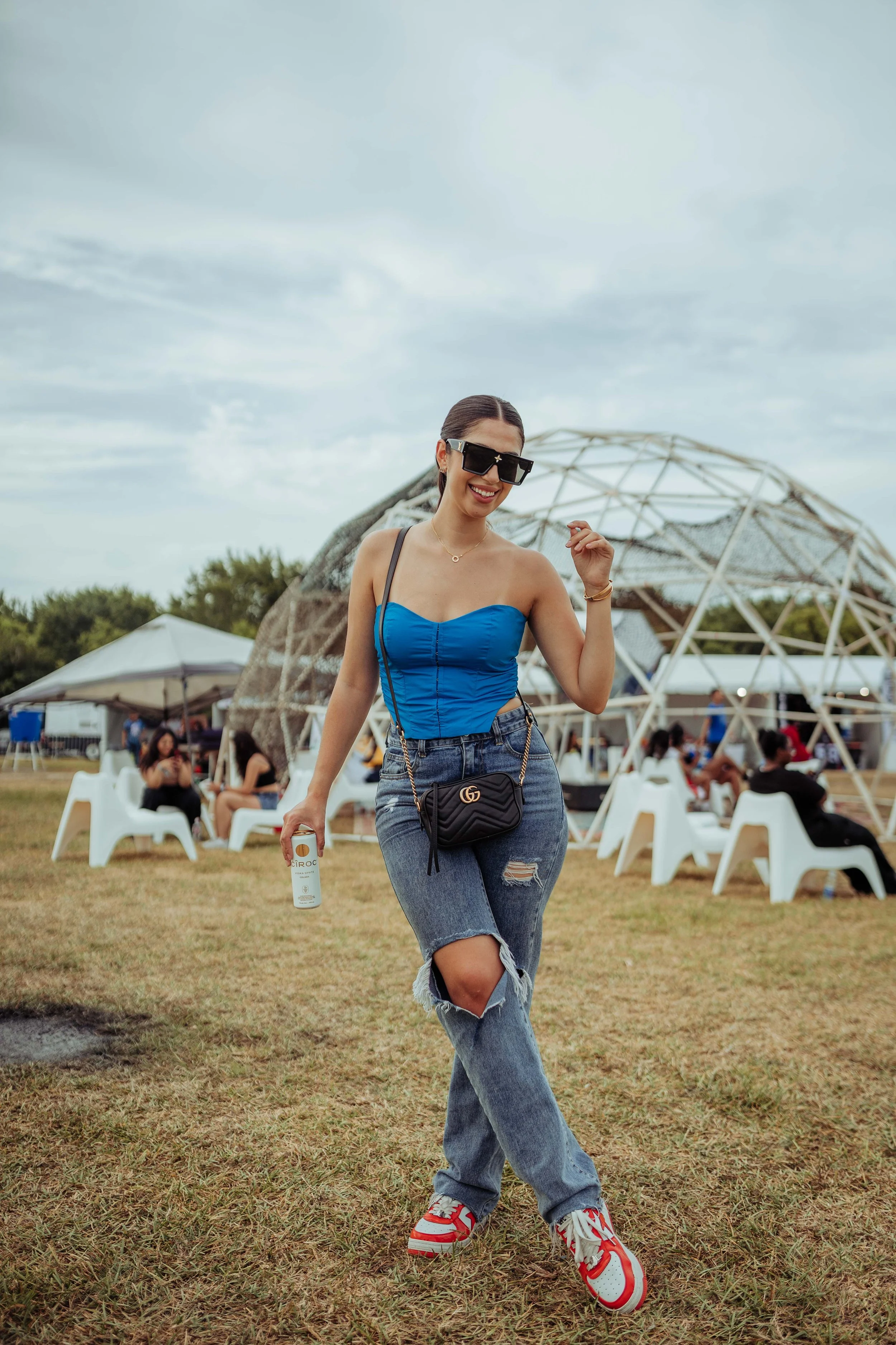 A young woman dancing outdoors at a festival, wearing sunglasses, a blue strapless top, ripped jeans, and red and white sneakers, holding a drink can and smiling.