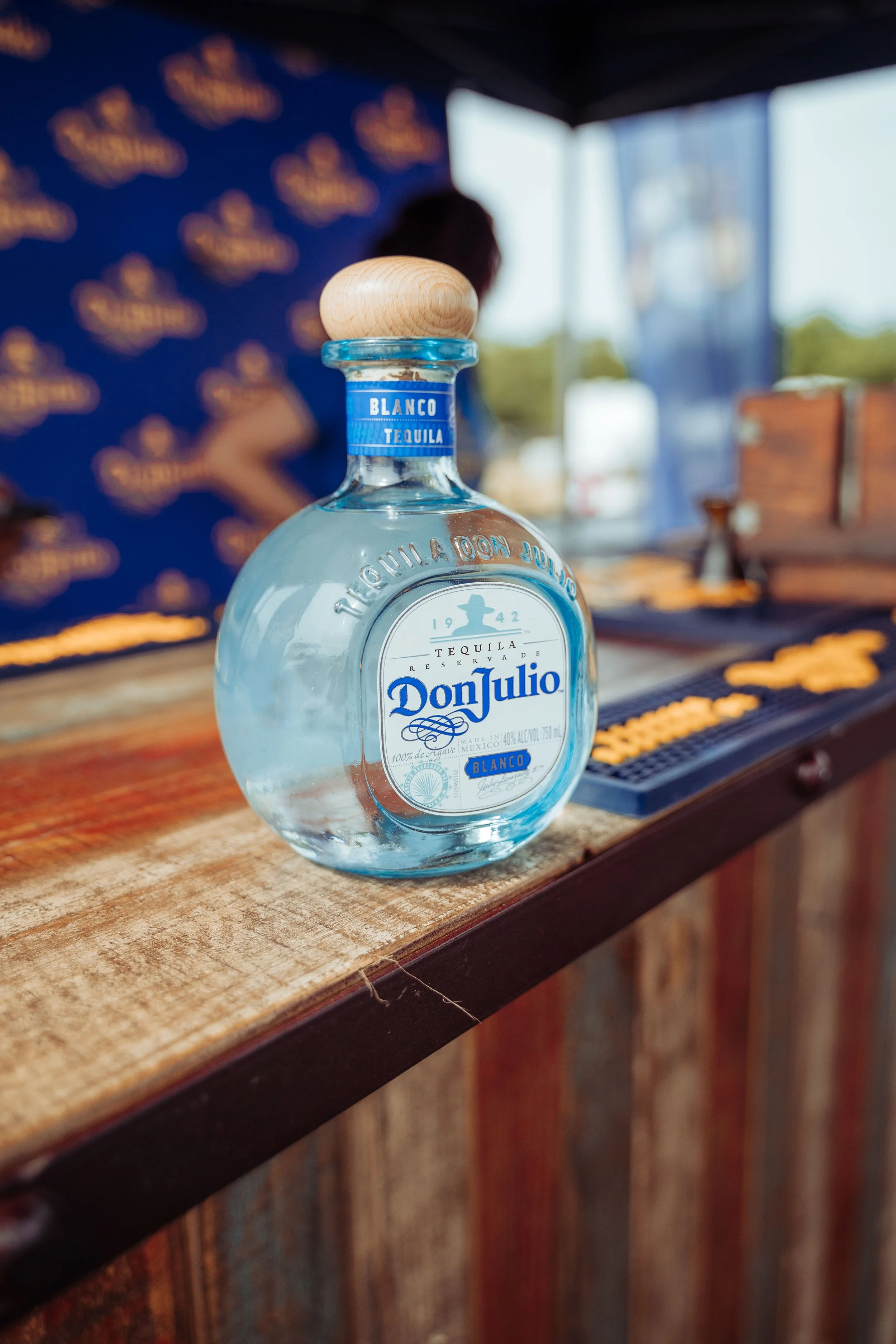 A clear glass bottle of Don Julio Blanco tequila on a wooden bar counter, with a blurred background of a person and a blue wall with yellow lettering.