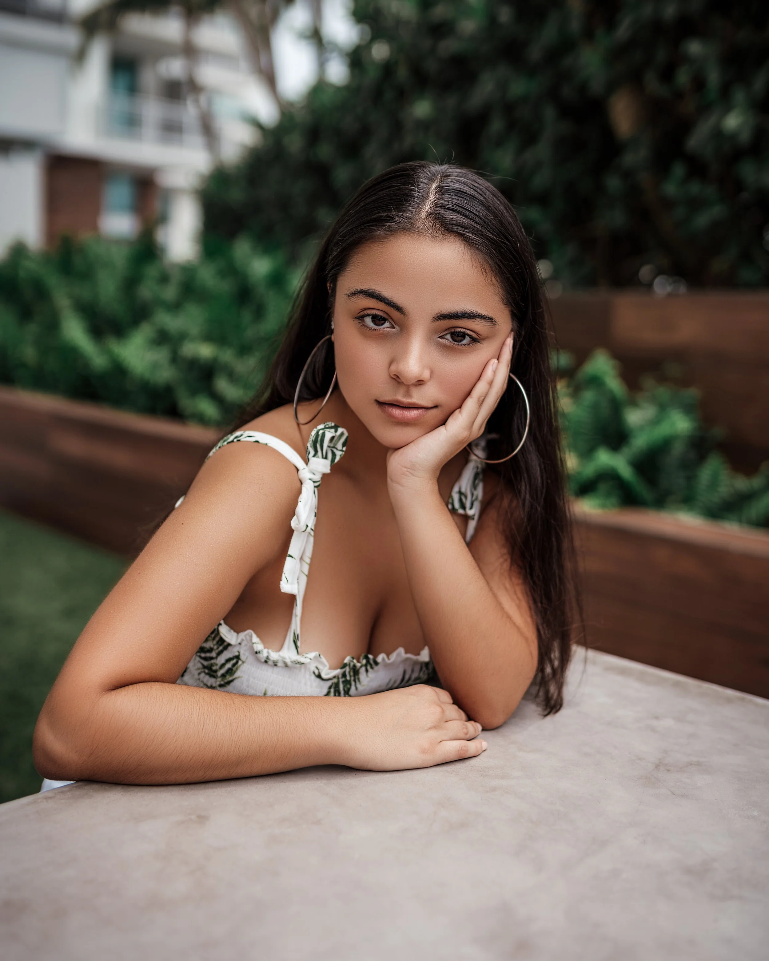 A young woman with long dark hair and hoop earrings resting her face on her hand, sitting at an outdoor table with greenery and buildings in the background.