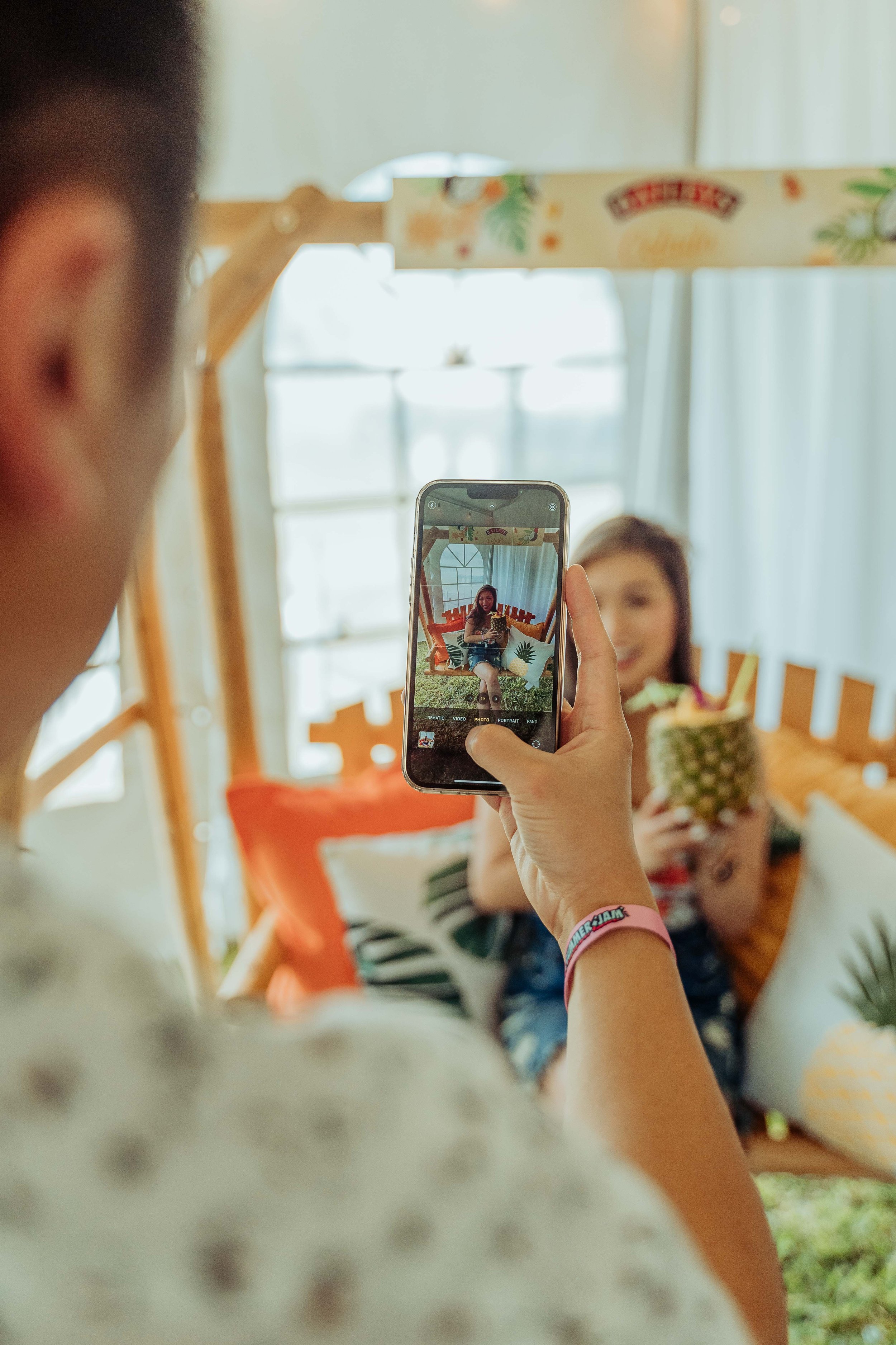 Person taking a photo of a woman sitting on a wooden bench, holding a pineapple drink, with colorful pillows and a bright window in the background.