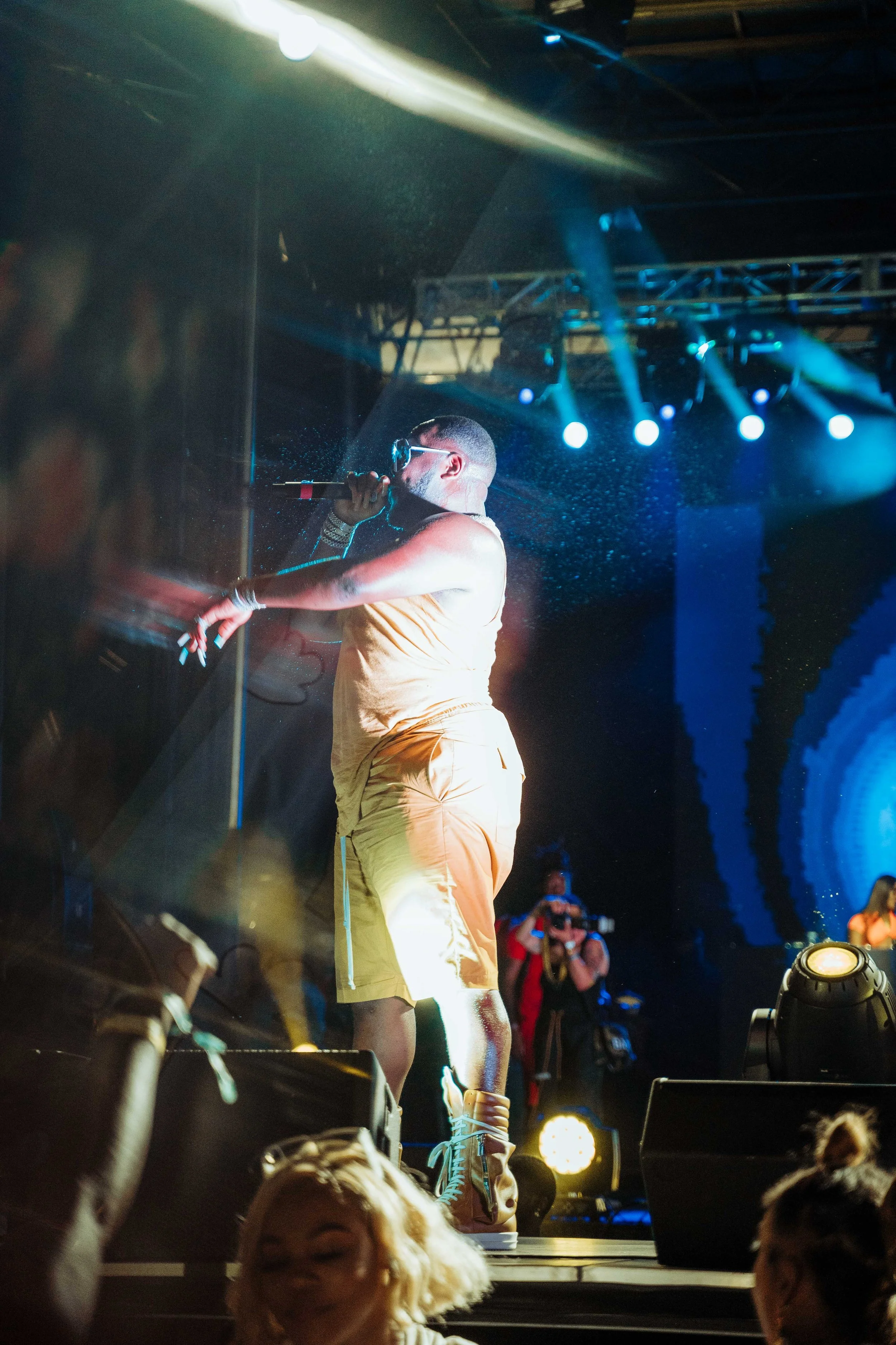 Performer on stage singing into a microphone during a concert, under colorful stage lights, with audience members in the foreground.