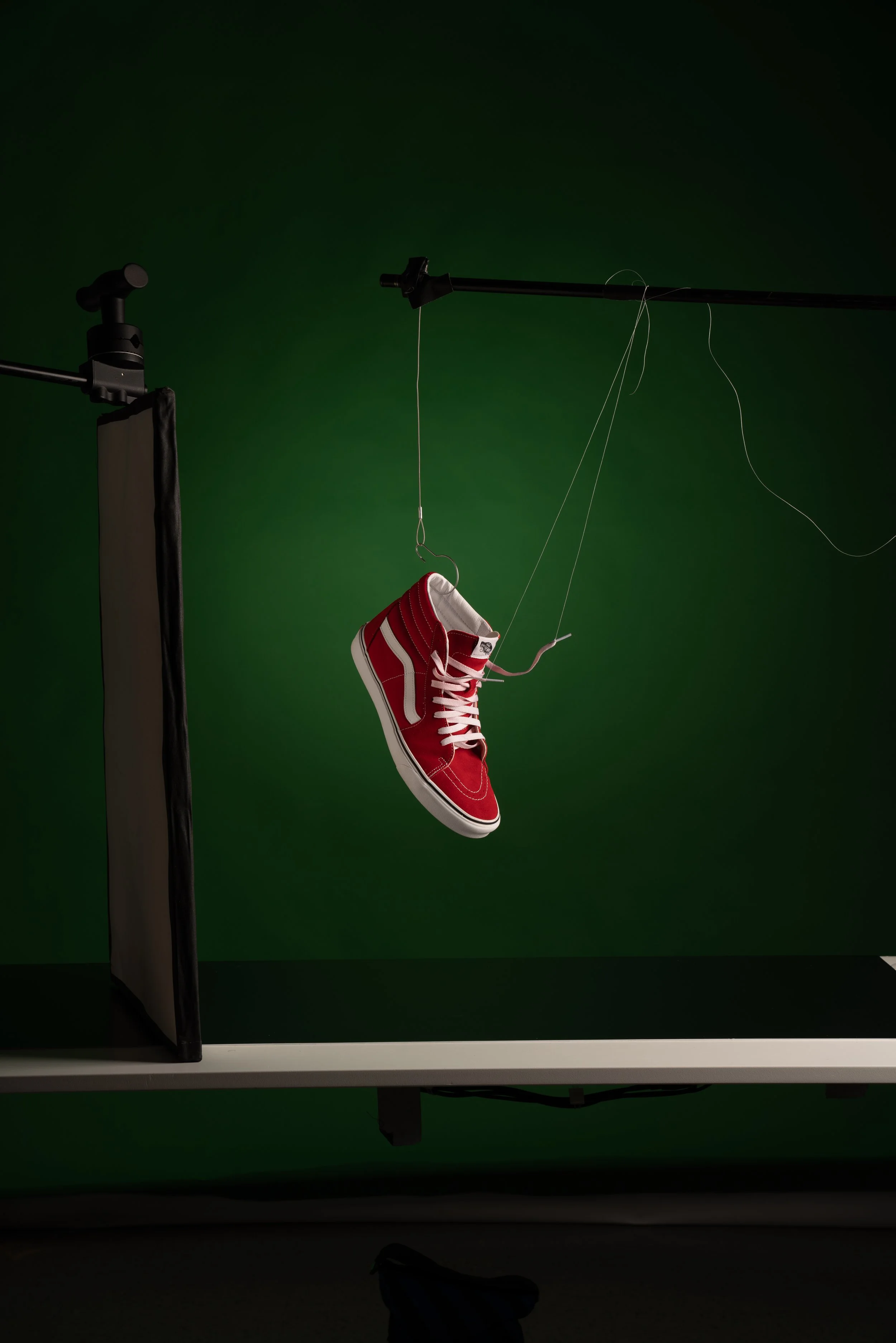 A red sneaker hanging from strings in front of a green screen during a photo shoot.
