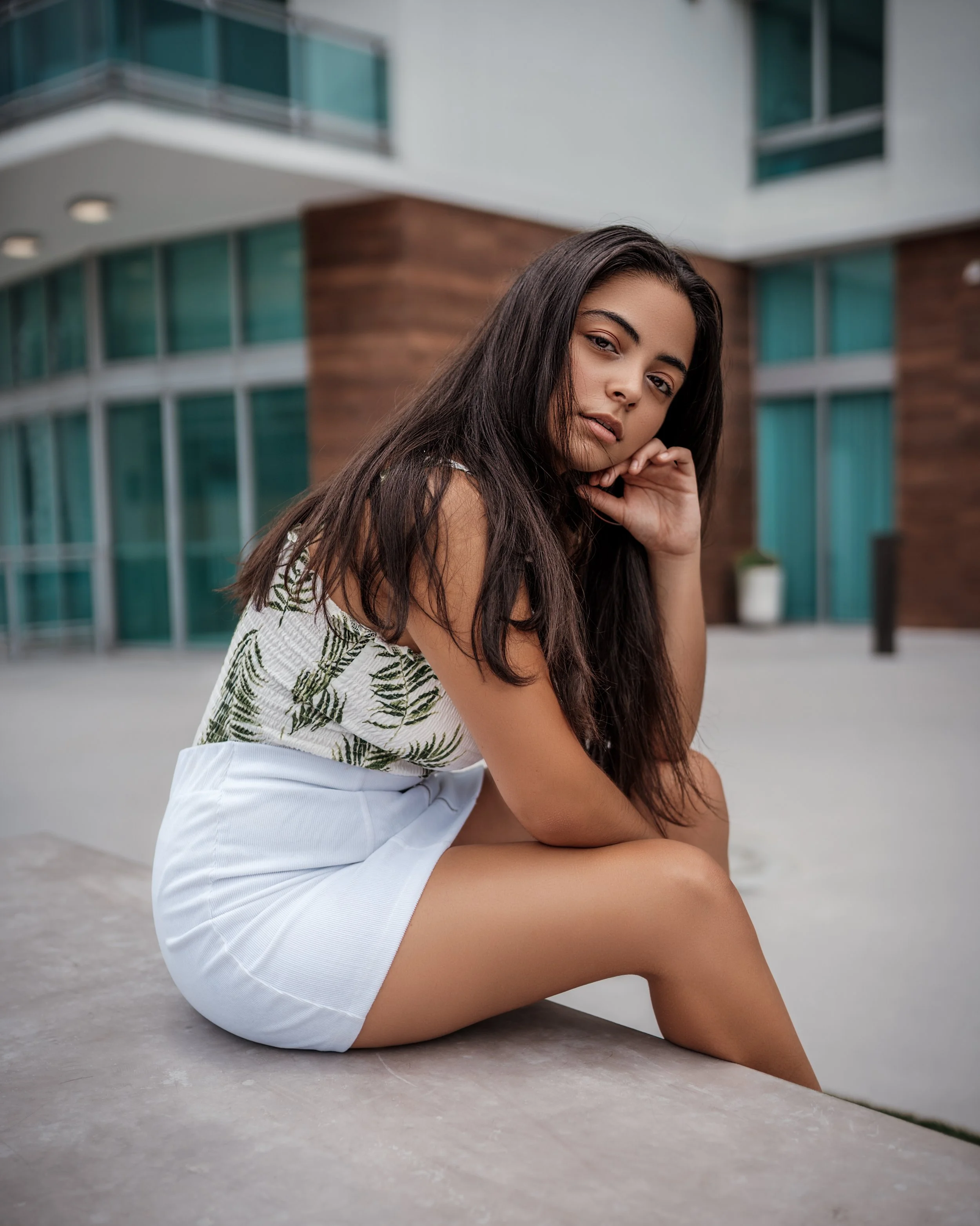 Young woman with long dark hair sitting outdoors near a modern building with glass windows, wearing a tropical print top and white skirt, looking at the camera with a contemplative expression.