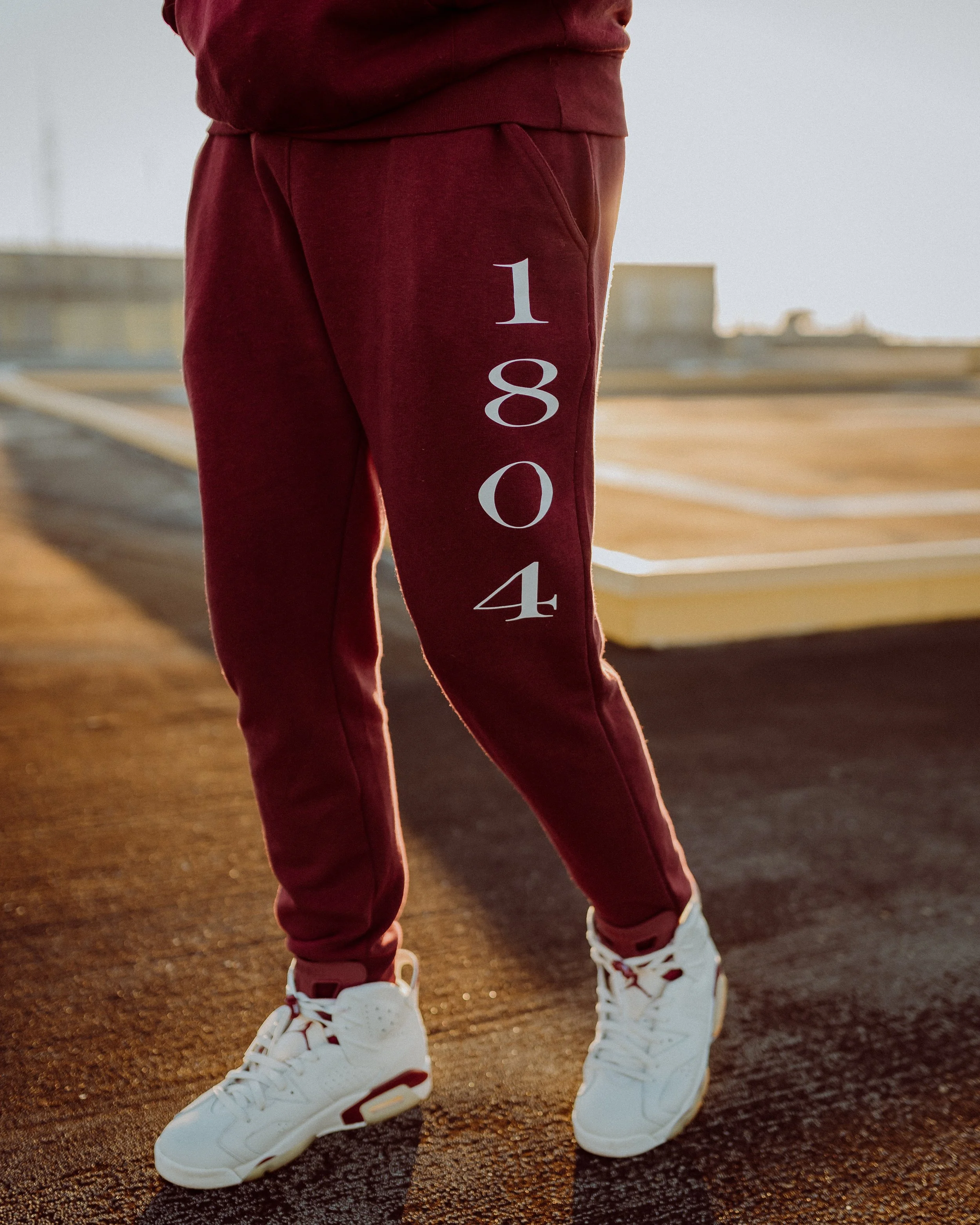 Person wearing maroon sweatpants with '1804' printed vertically on the left leg and white sneakers standing on a basketball court.
