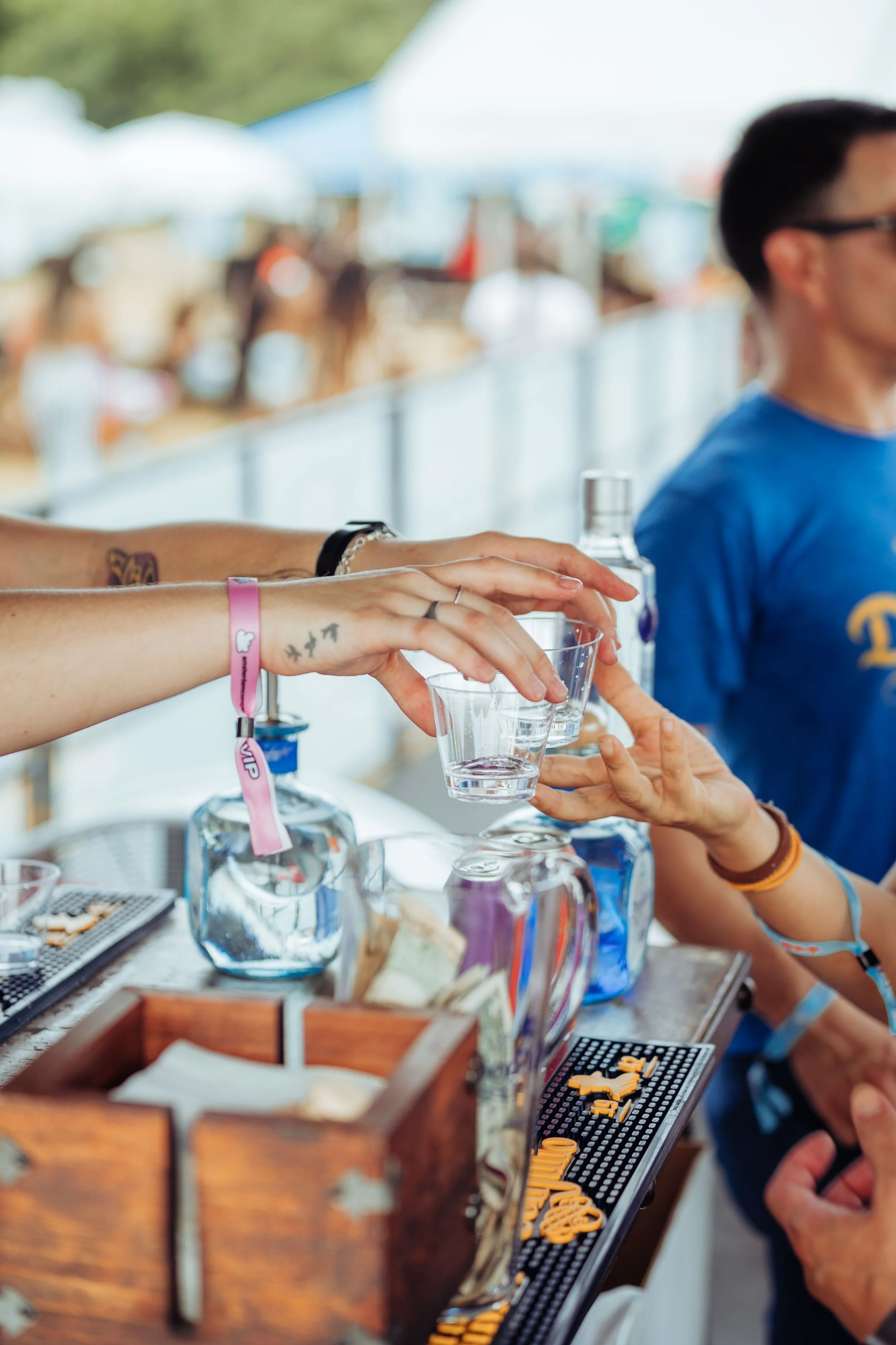 Person with tattoos and bracelet handing a plastic cup to another person at an outdoor bar, with beer tap and bar supplies visible.