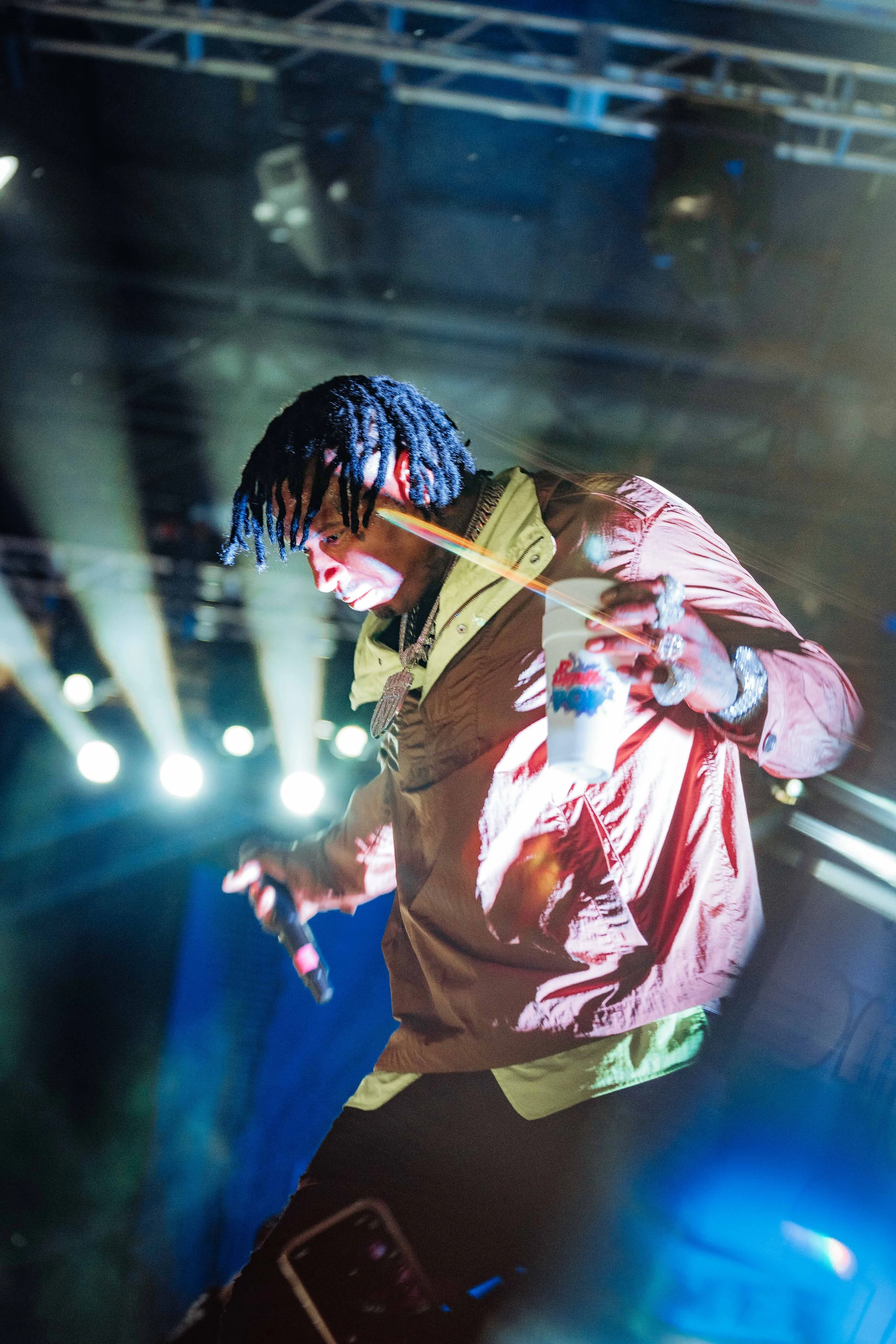 A performer with black and blue dreadlocks on stage holding a microphone and a cup, illuminated by stage lights.